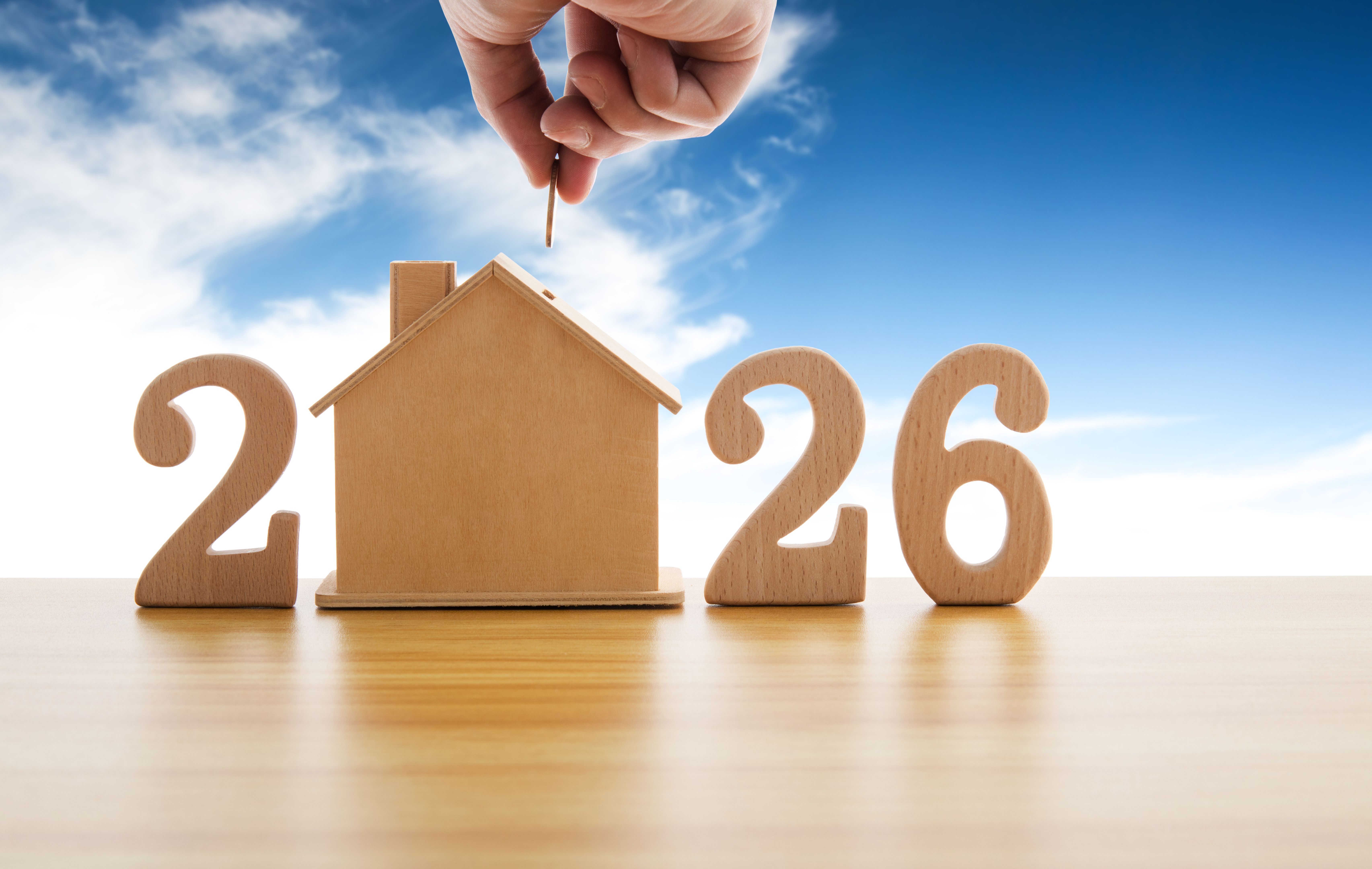 Closeup of hand putting coin in house piggy bank with wooden number 2026 against blue sky