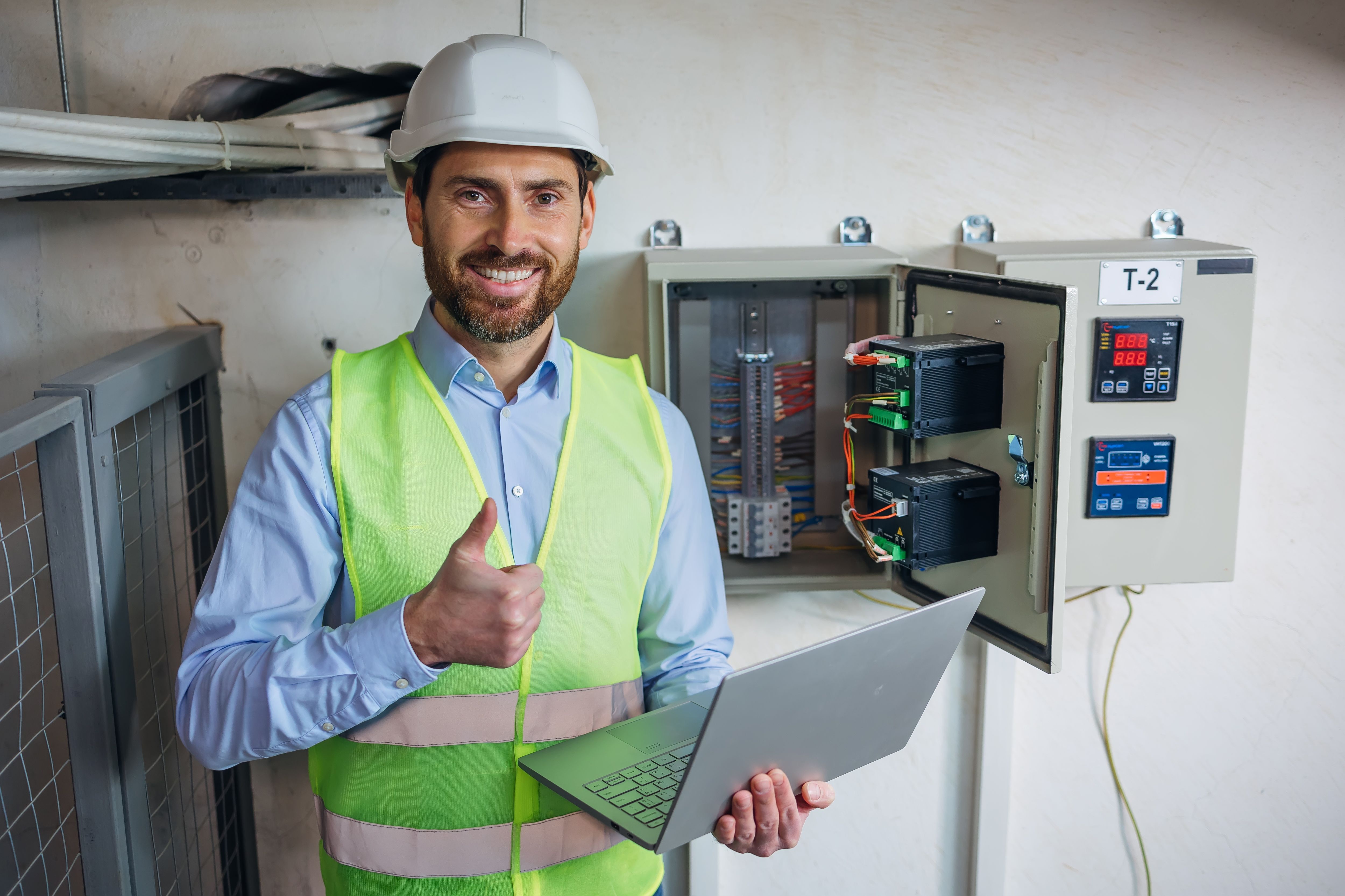 Professional inspector electrician technician engineer in safety uniform holding laptop checking power voltage electrical system showing thumbs up and Smiling at industrial factory