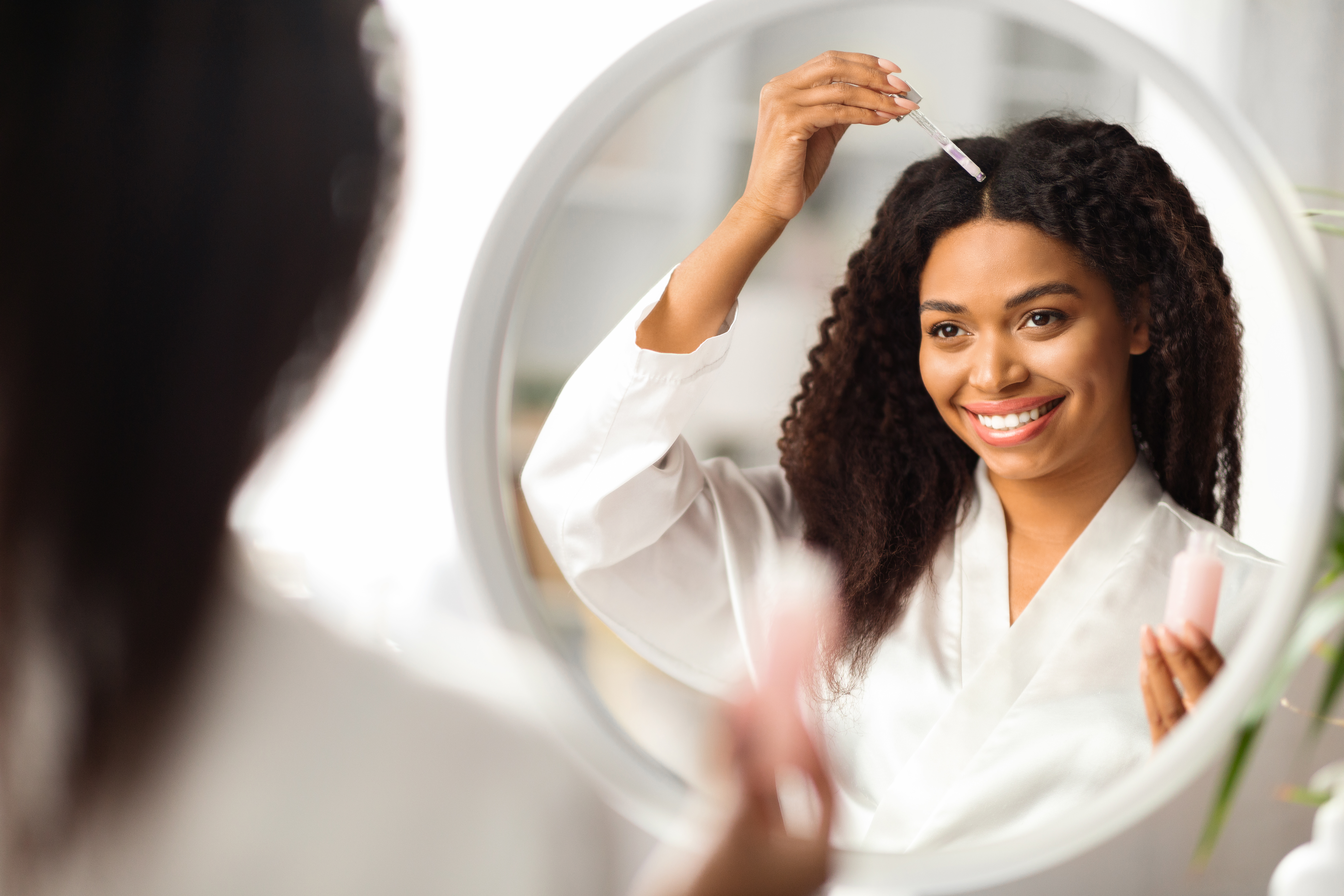 Smiling Black Woman Applying Serum For Hair Repair While Standing Near Mirror Smiling Black Woman Applying Serum For Hair Repair While Standing Near Mirror