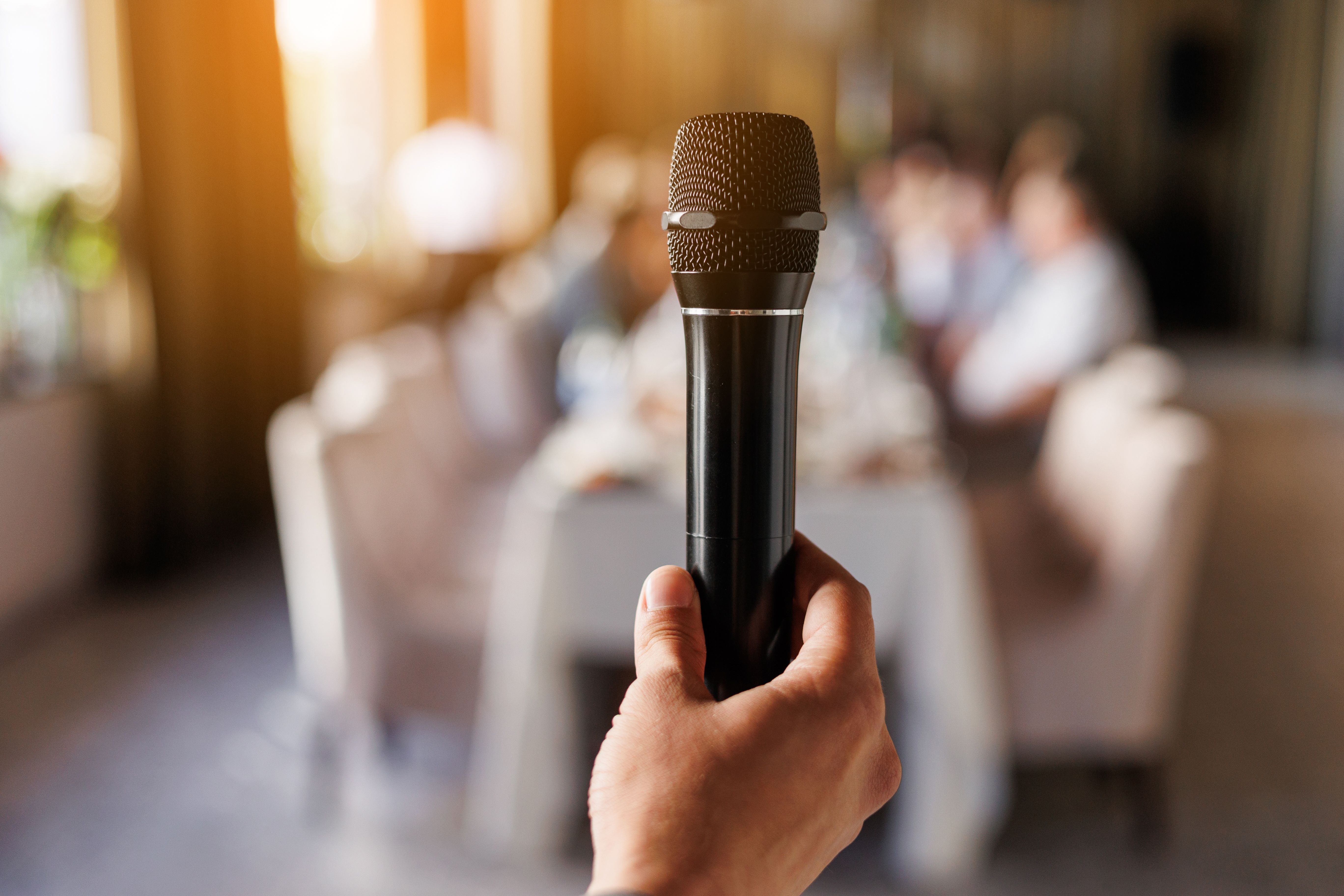 Unrecognisable male hand holds black microphone, master of ceremony or showman speech to audience in event. On background sit people guests by the table. Party and festive events concept.