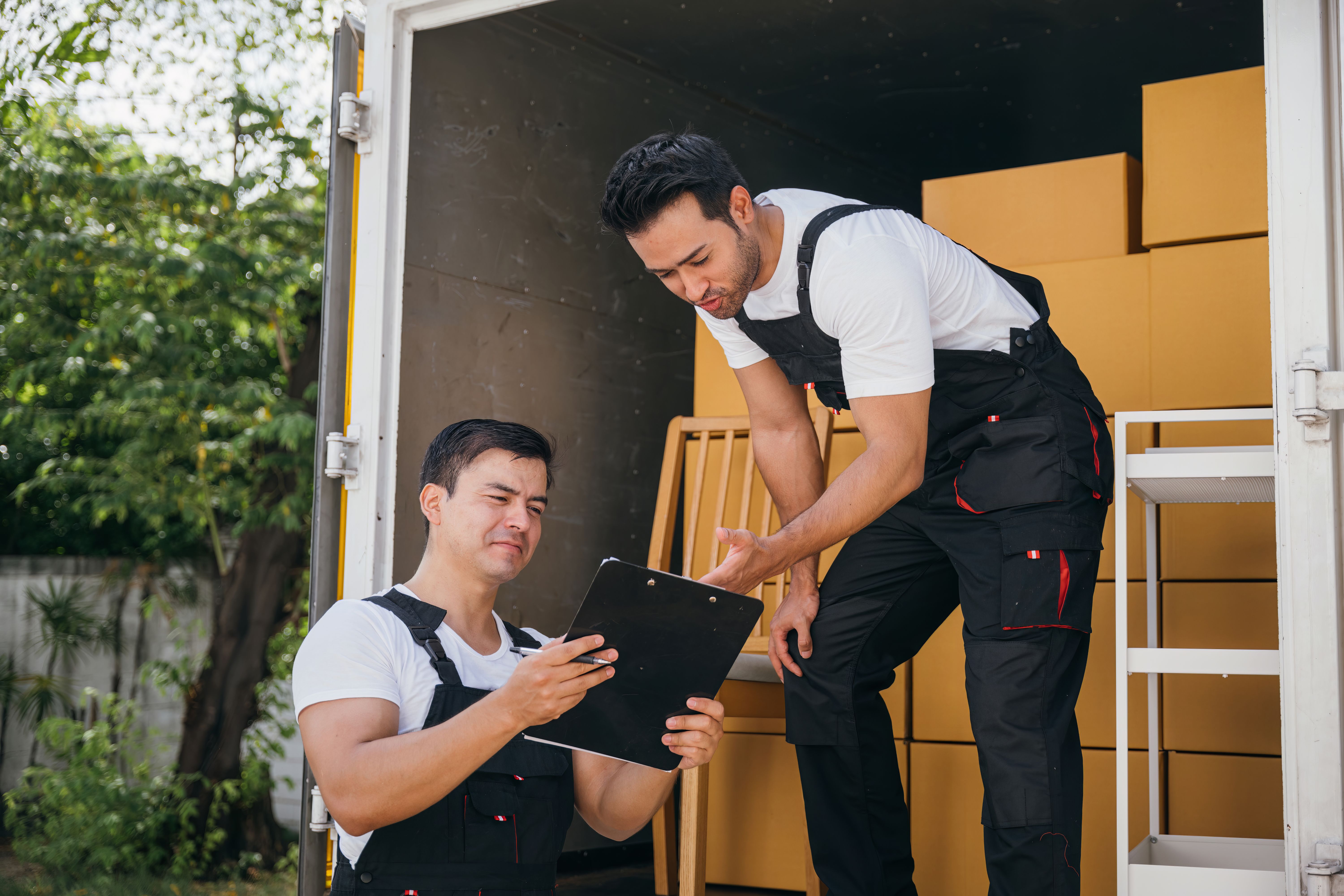 Delivery men in uniform unload boxes inspecting shipment with a checklist and clipboard at the truck's back. Expert movers guarantee efficient moving and reliable service. Moving day concept Delivery men in uniform unload boxes inspecting shipment with a checklist and clipboard at the truck's back. Expert movers guarantee efficient moving and reliable service. Moving day concept