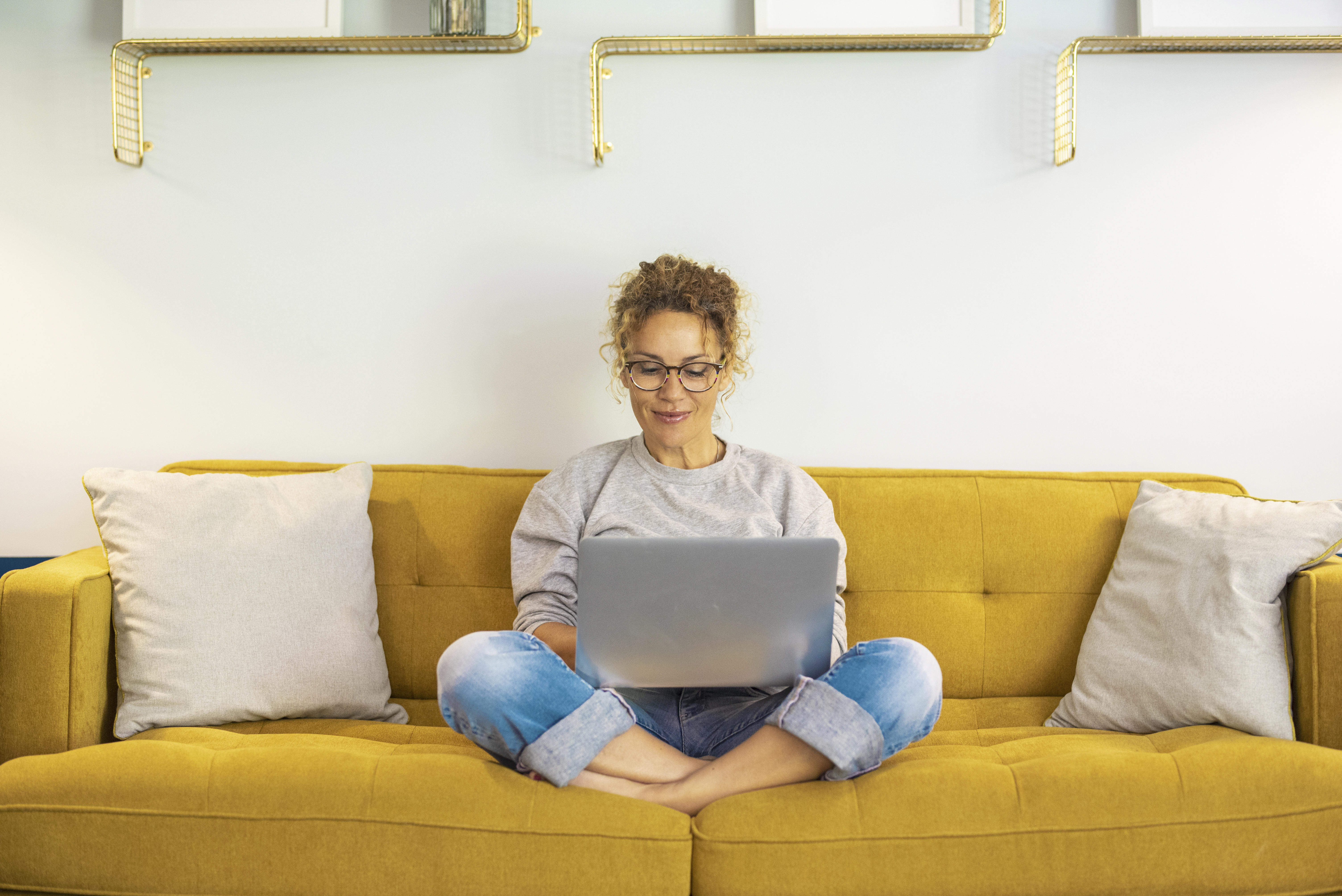 One woman smiling and using laptop computer at home sitting comfortably on a yellow sofa in living room. Smart working female people notebook. Surfing the web. Enjoying technology and connection