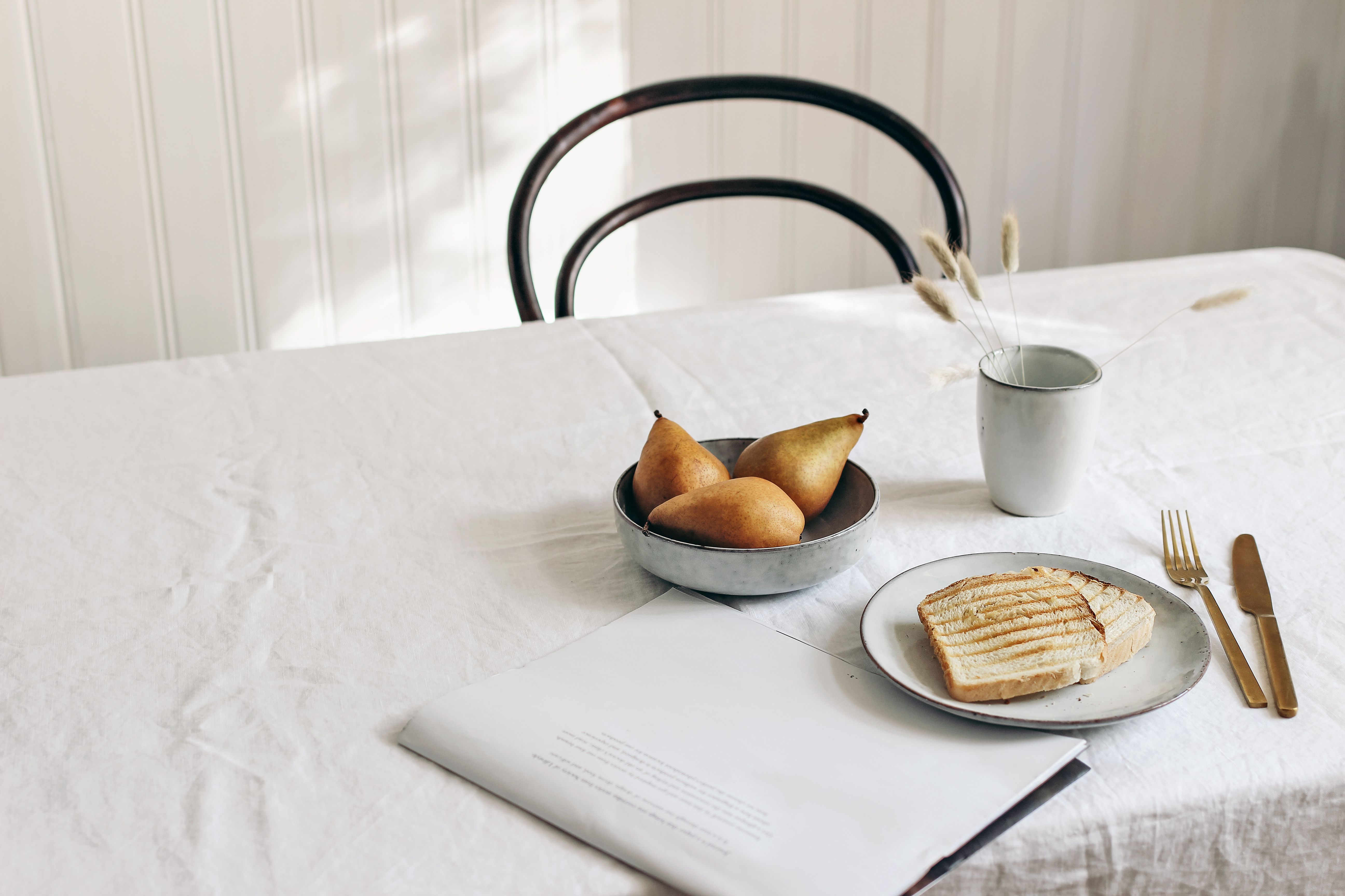 Morning breakfast with pears fruit composition. Bread toast, folded newspapers and dry grass in mug on white linen table cloth. Minimal Scandinavian interior, dinning room in sunlight. Nordic kitchen.