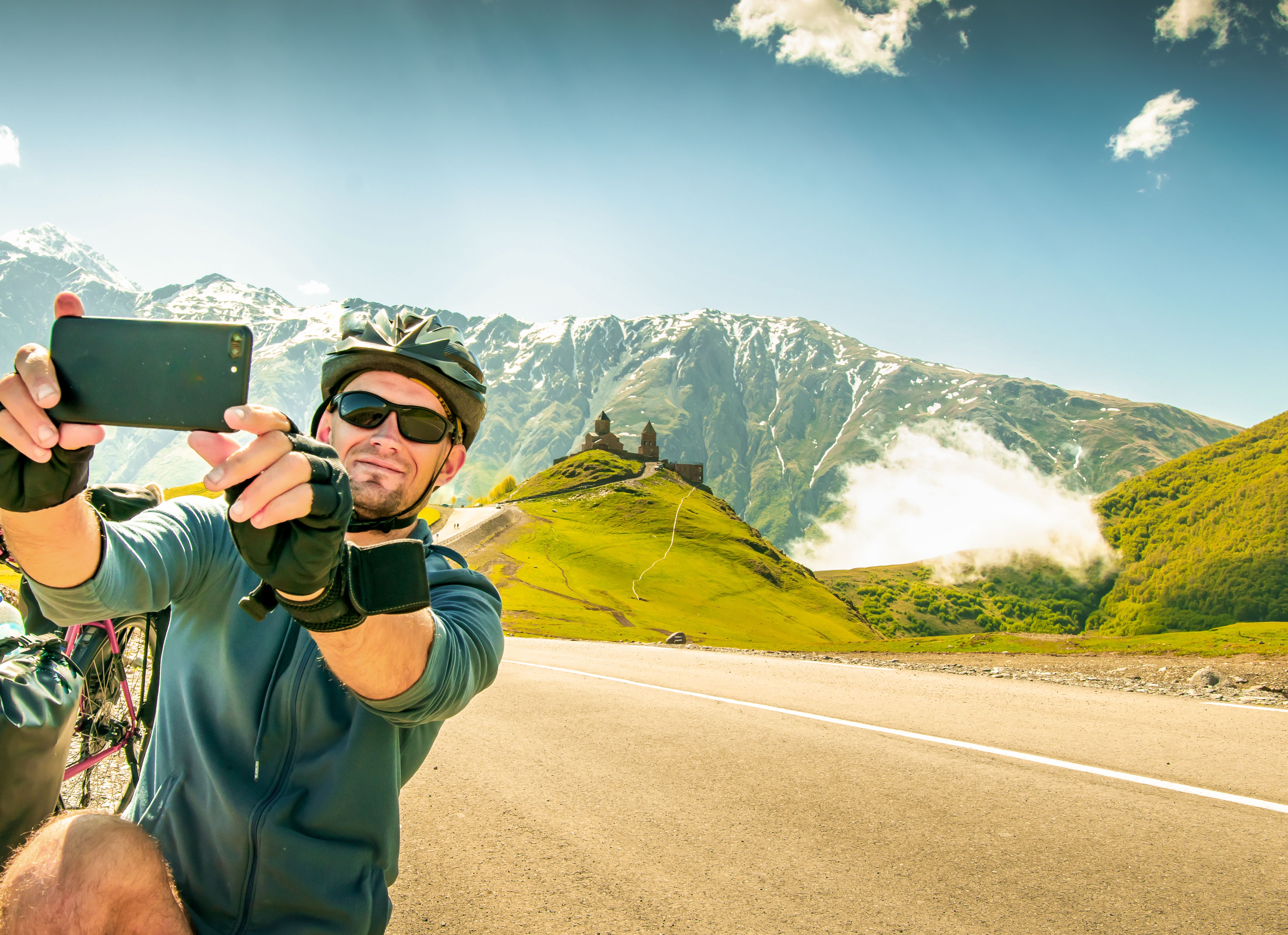 Young caucasian cyclist by touring bicycle make self-portrait outdoors while bicycle touring on hilltop viewpoint. Gergeti trinity church. Kazbegi national park