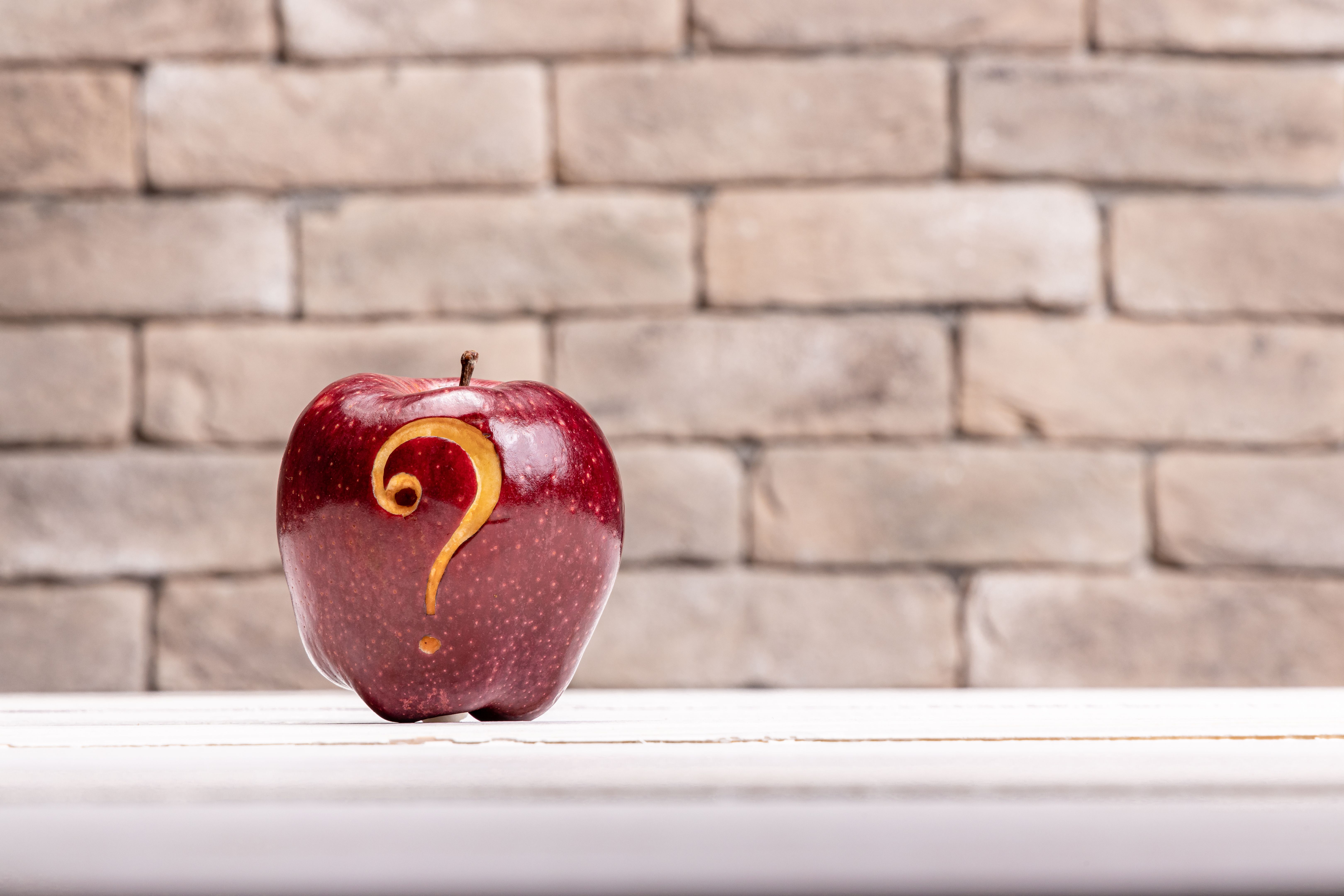 Close-up of red shiny apple with carved question mark sigh on white light surface with brick wall background- creative stock photo