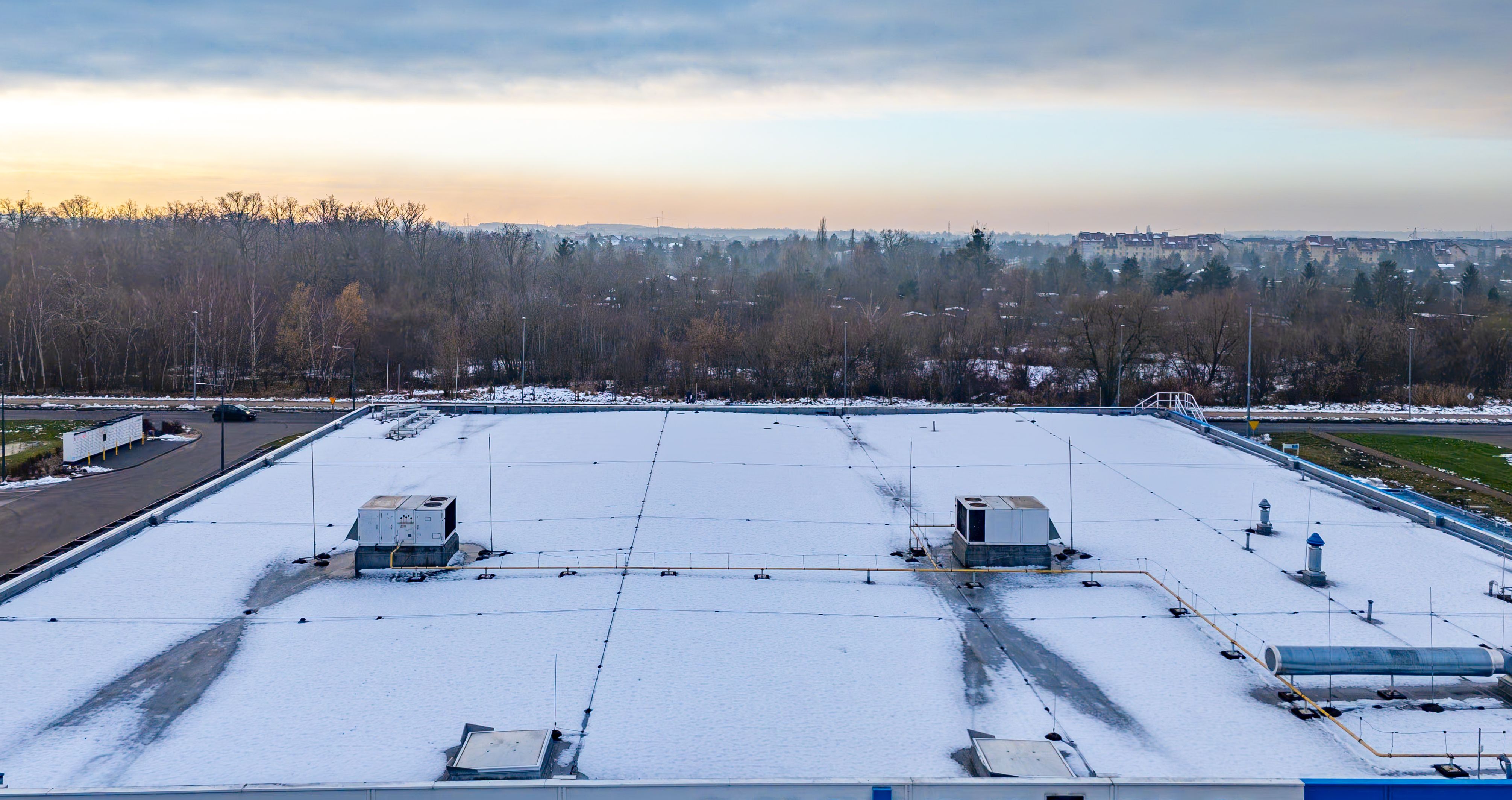 Aerial view of a snow-covered industrial hall roof with ventilation units, technical installations, surrounding woodland and distant residential buildings