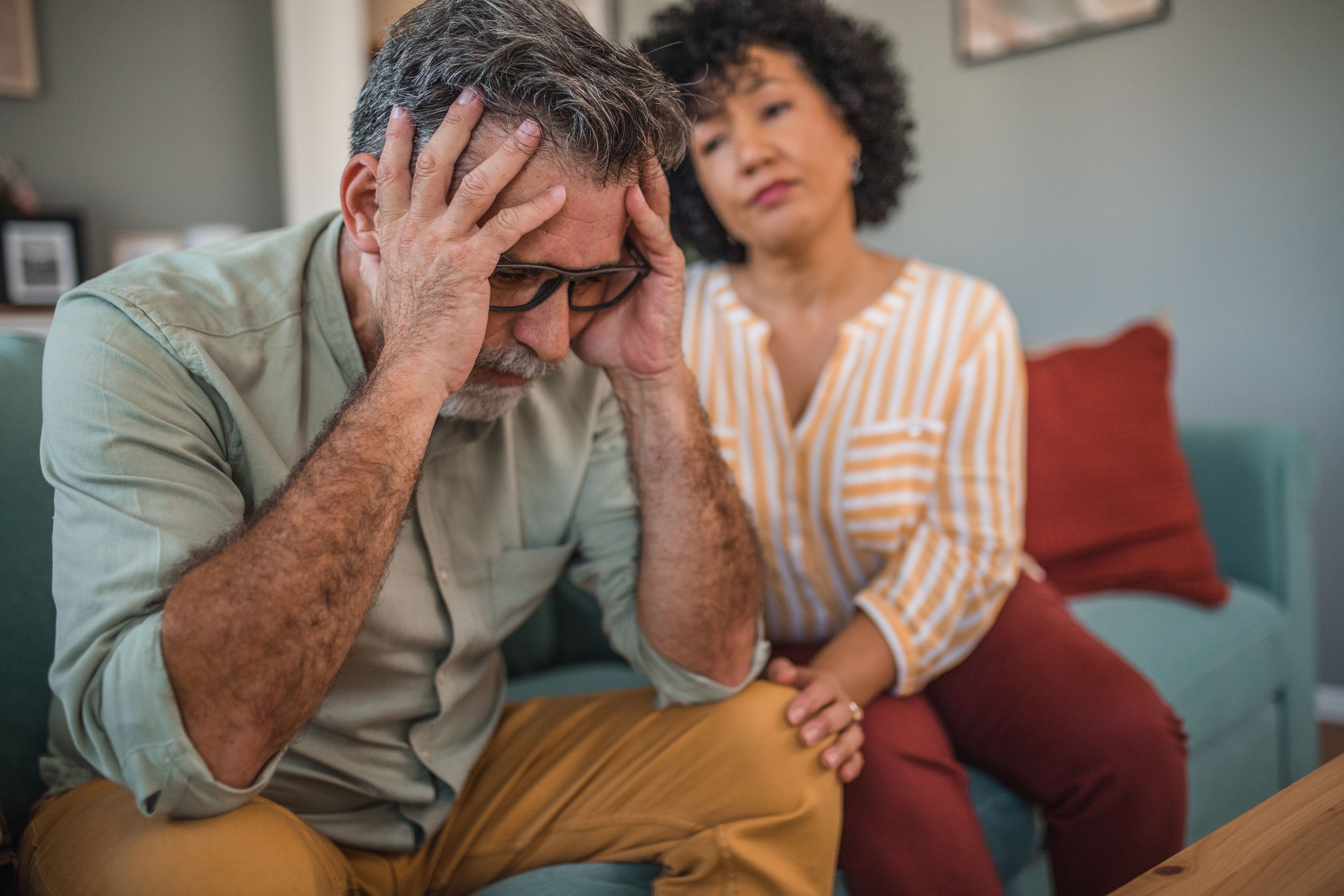 A middle-aged man has a severe headache, his mixed race woman comforts him. They are sitting in the living room A middle-aged man has a severe headache, his mixed race woman comforts him. They are sitting in the living room