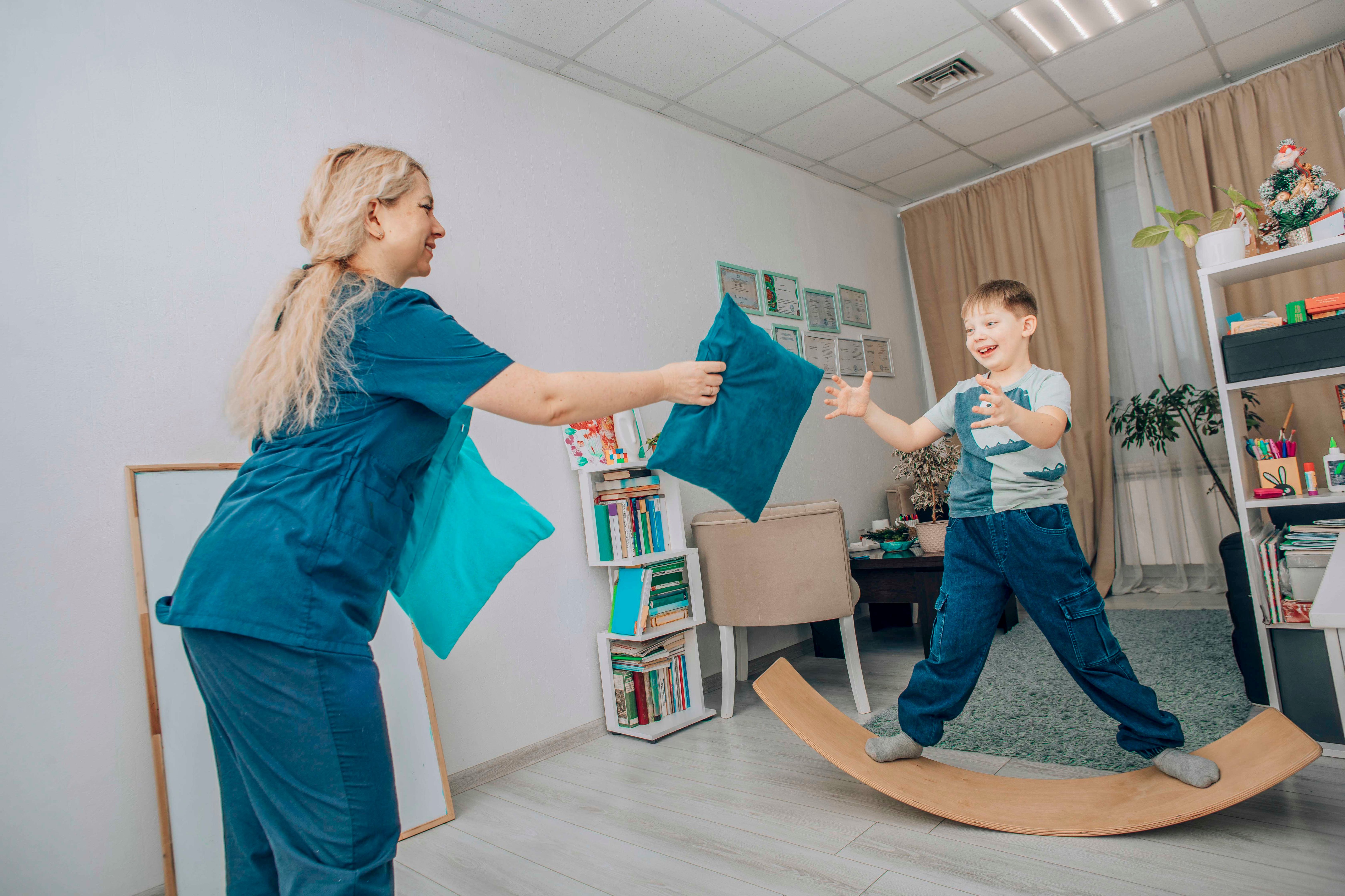 Smiling Boy On Wooden Board, Playing With Therapist In Bright Room, Fun Pillow, Physical Therapy Smiling Boy On Wooden Board, Playing With Therapist In Bright Room, Fun Pillow, Physical Therapy