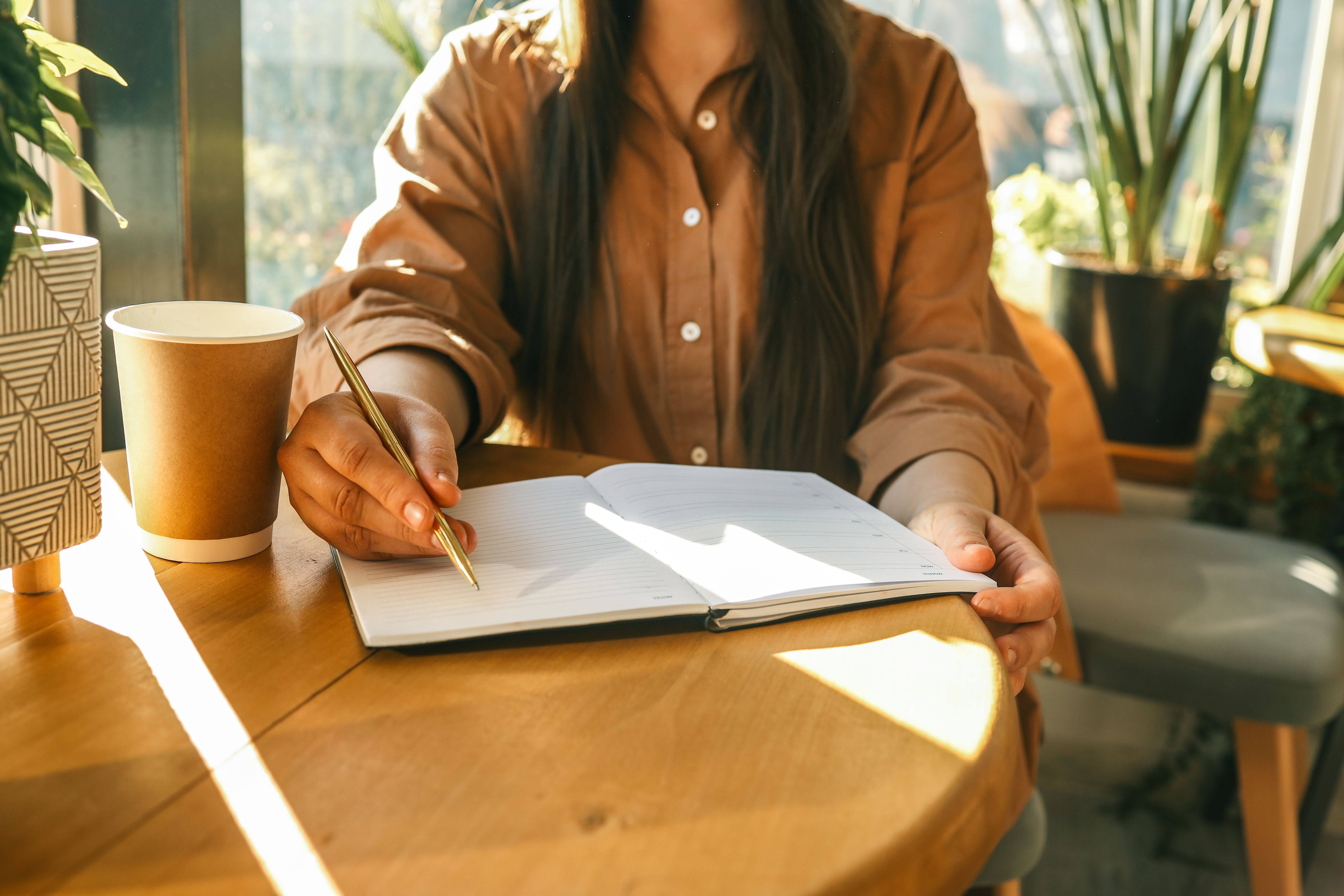 Woman writing in open planner at sunny cafÃ© table with takeaway coffee cup and houseplants, wearing brown shirt, natural light coming through large window