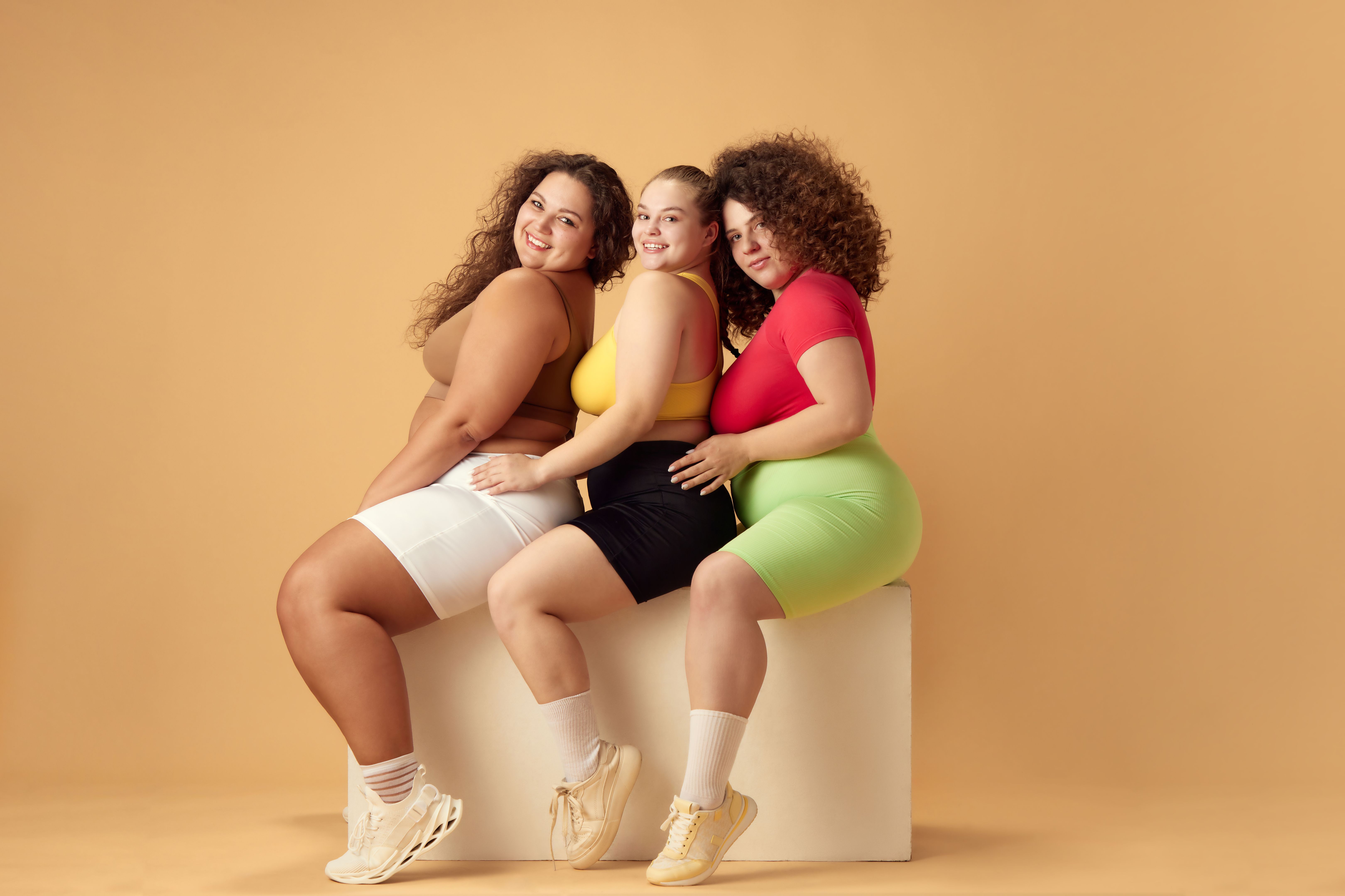 Friends, beautiful women with oversized bodies, wearing sportswear, posing over beige studio background Friends, beautiful women with oversized bodies, wearing sportswear, posing over beige studio background