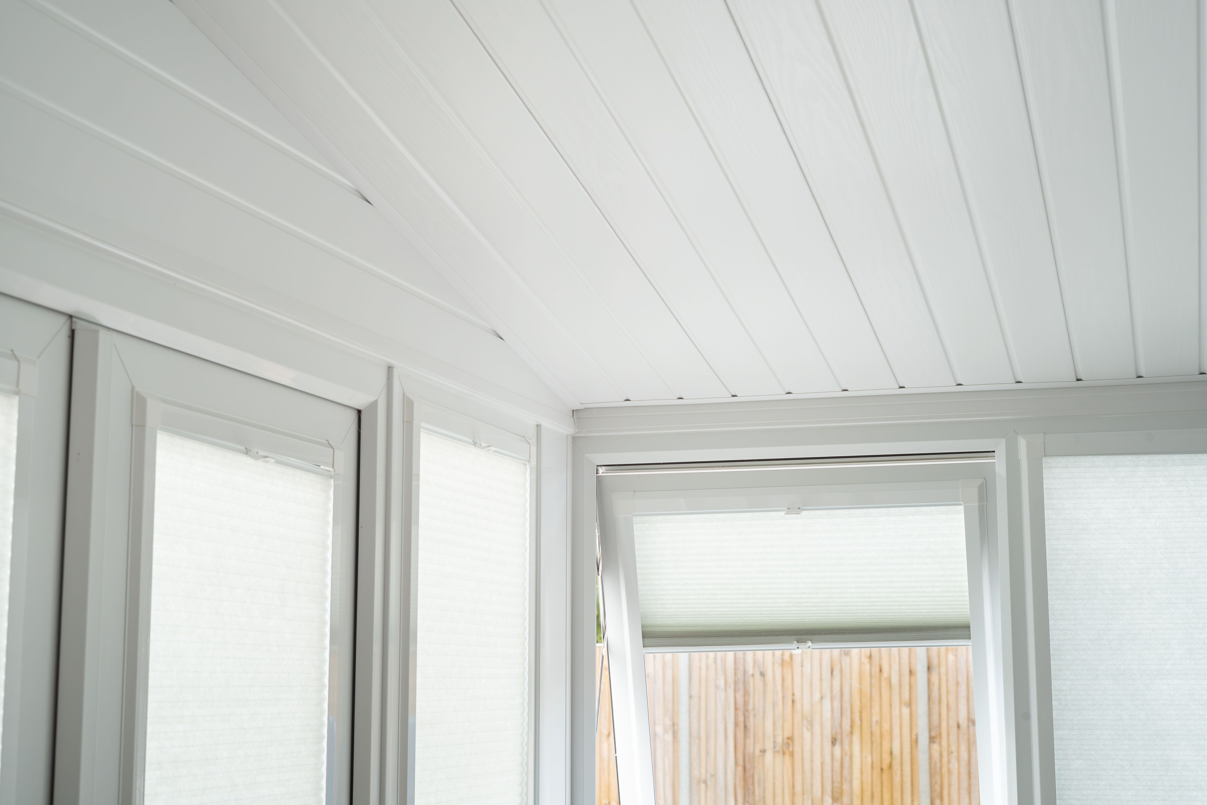 An insulated white conservatory, sunroom roof, sometimes called a warm roof, constructed with a plank or beam wood texture design and window blinds.