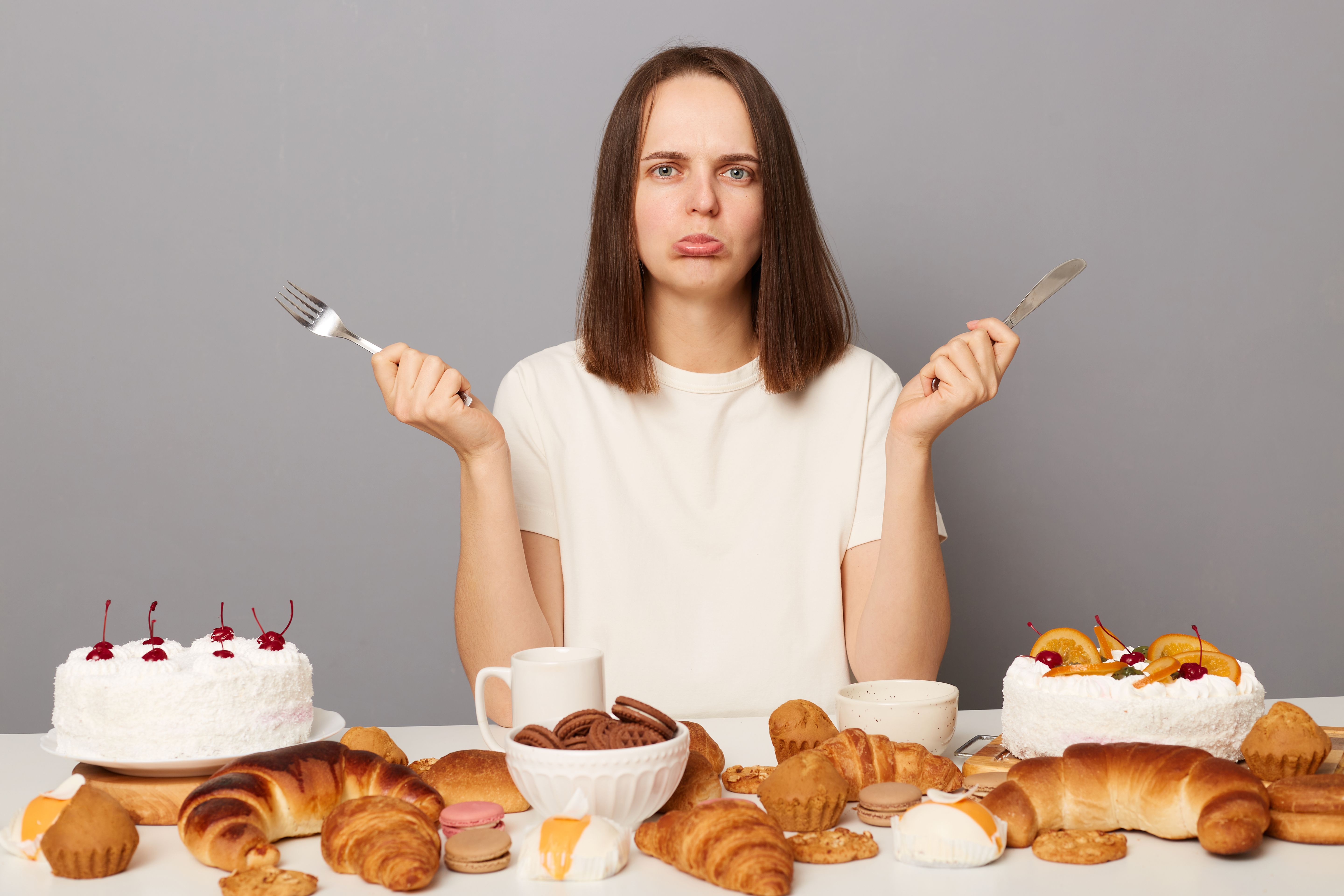 Portrait of sad stressed Caucasian woman with brown hair sitting at table isolated over gray background, looking at camera with pout lips, being upset, keeps diet, wants sweet bakery. Portrait of sad stressed Caucasian woman with brown hair sitting at table isolated over gray background, looking at camera with pout lips, being upset, keeps diet, wants sweet bakery.