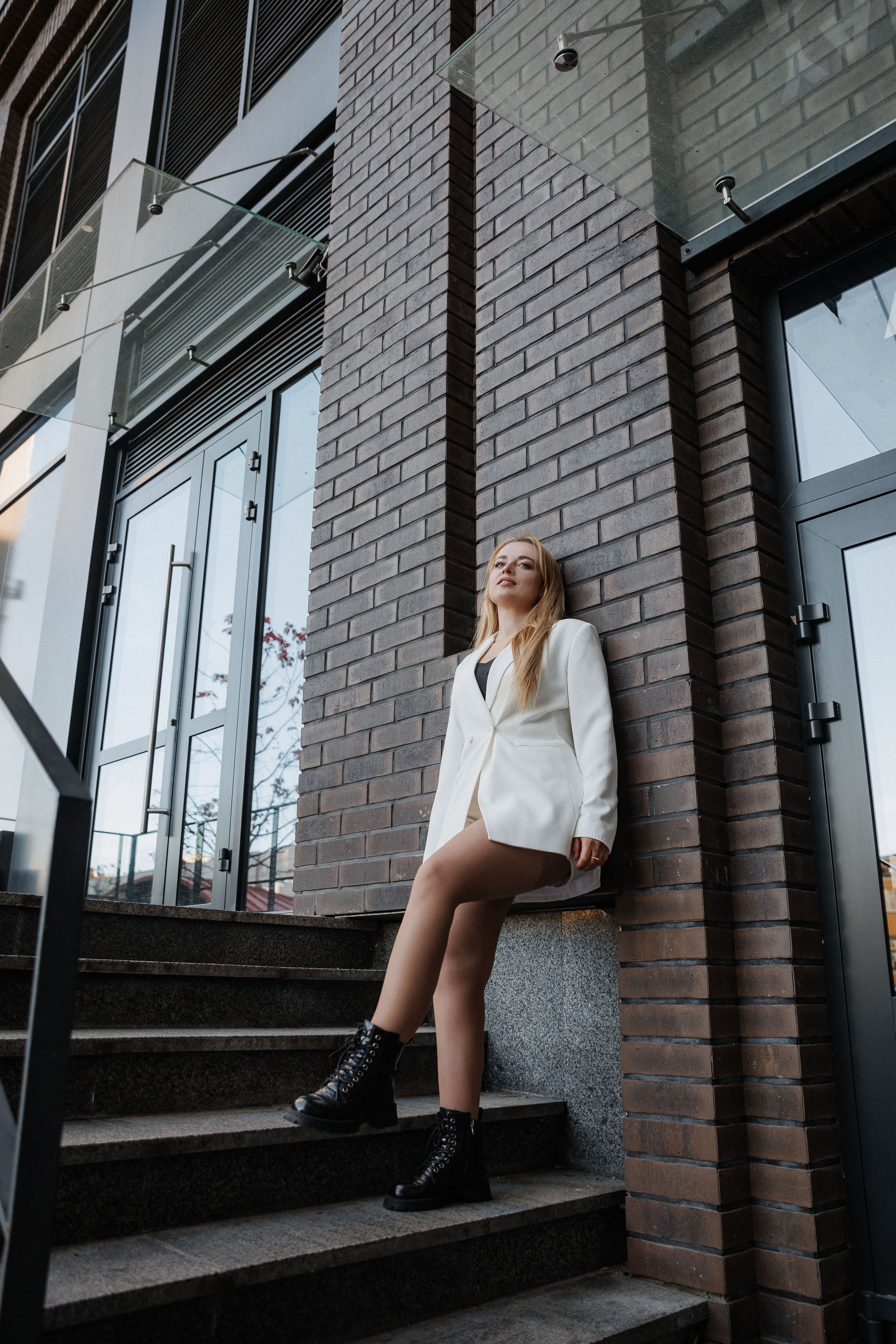 Blond stylish woman at oversized white jacket posing at the railing and steps on the city background Blond stylish woman at oversized white jacket posing at the railing and steps on the city background