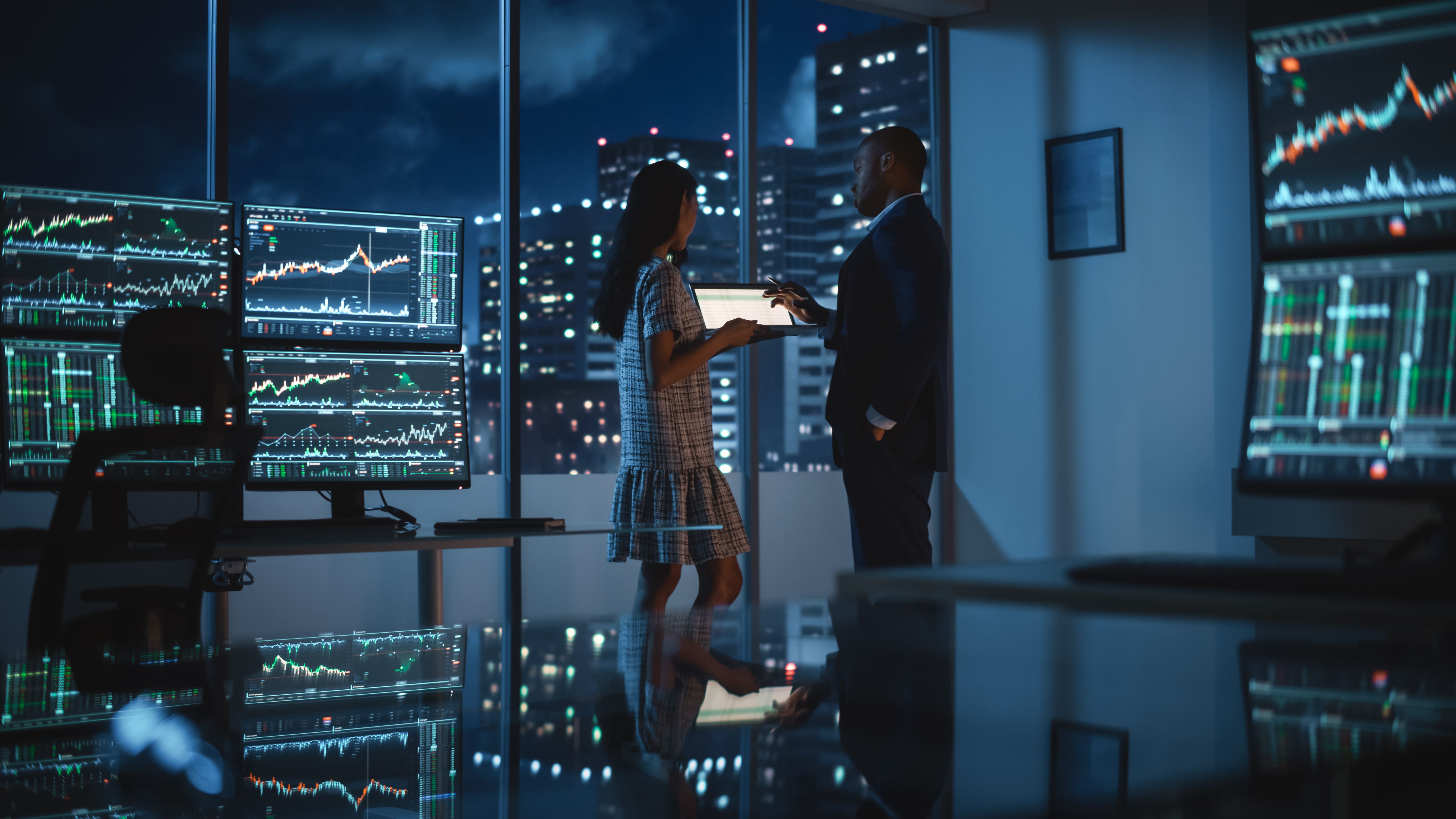 Financial Analyst Talking to Investment Banker in Late Evening at Work. Chatting About Real-Time Stock Chart Data on Tablet Computer. Businesspeople Have a Meeting in Broker Agency City Office.