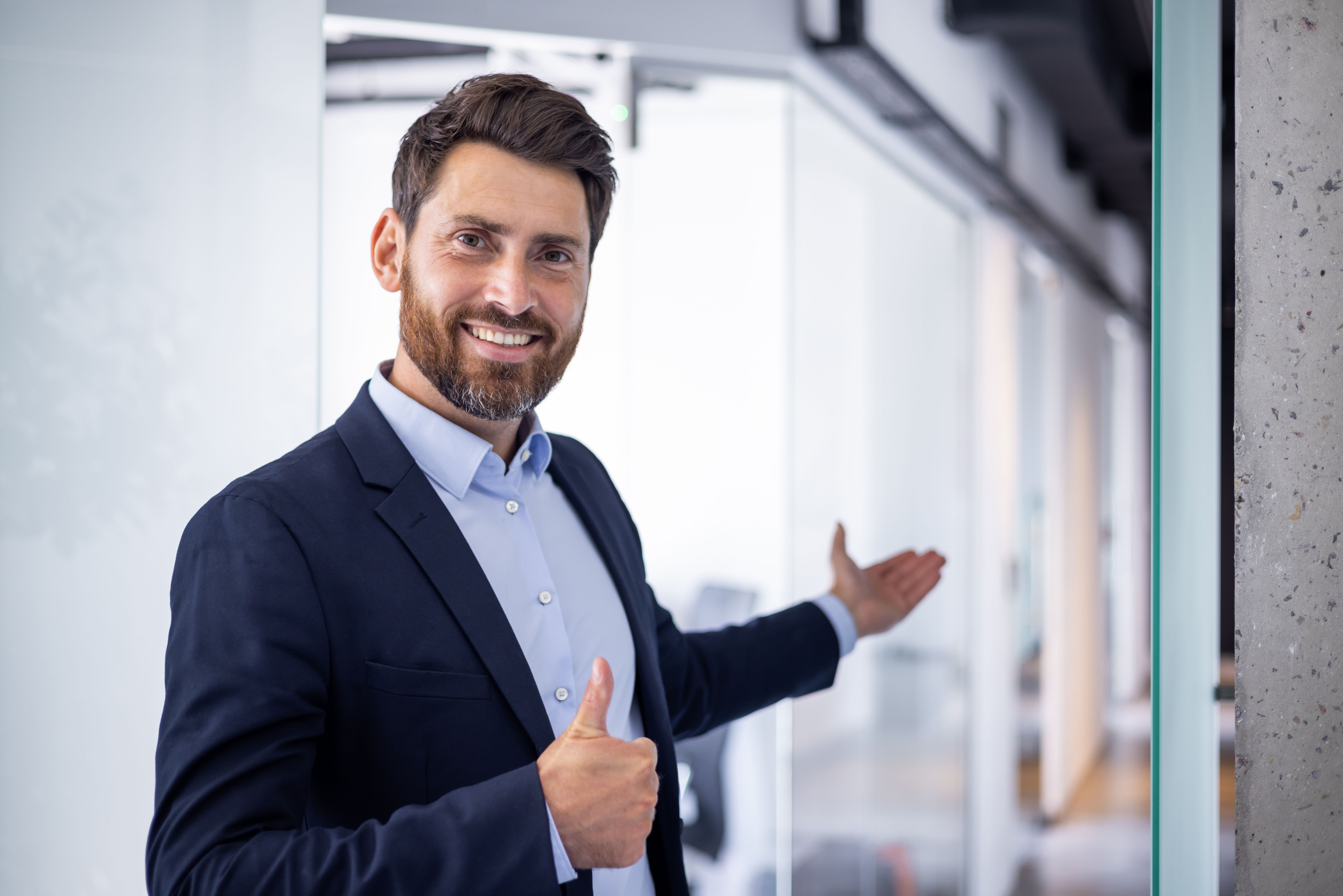 A young smiling male businessman, the founder of the company stands in a suit and invites to the office with a hand gesture of welcome and shows the super finger