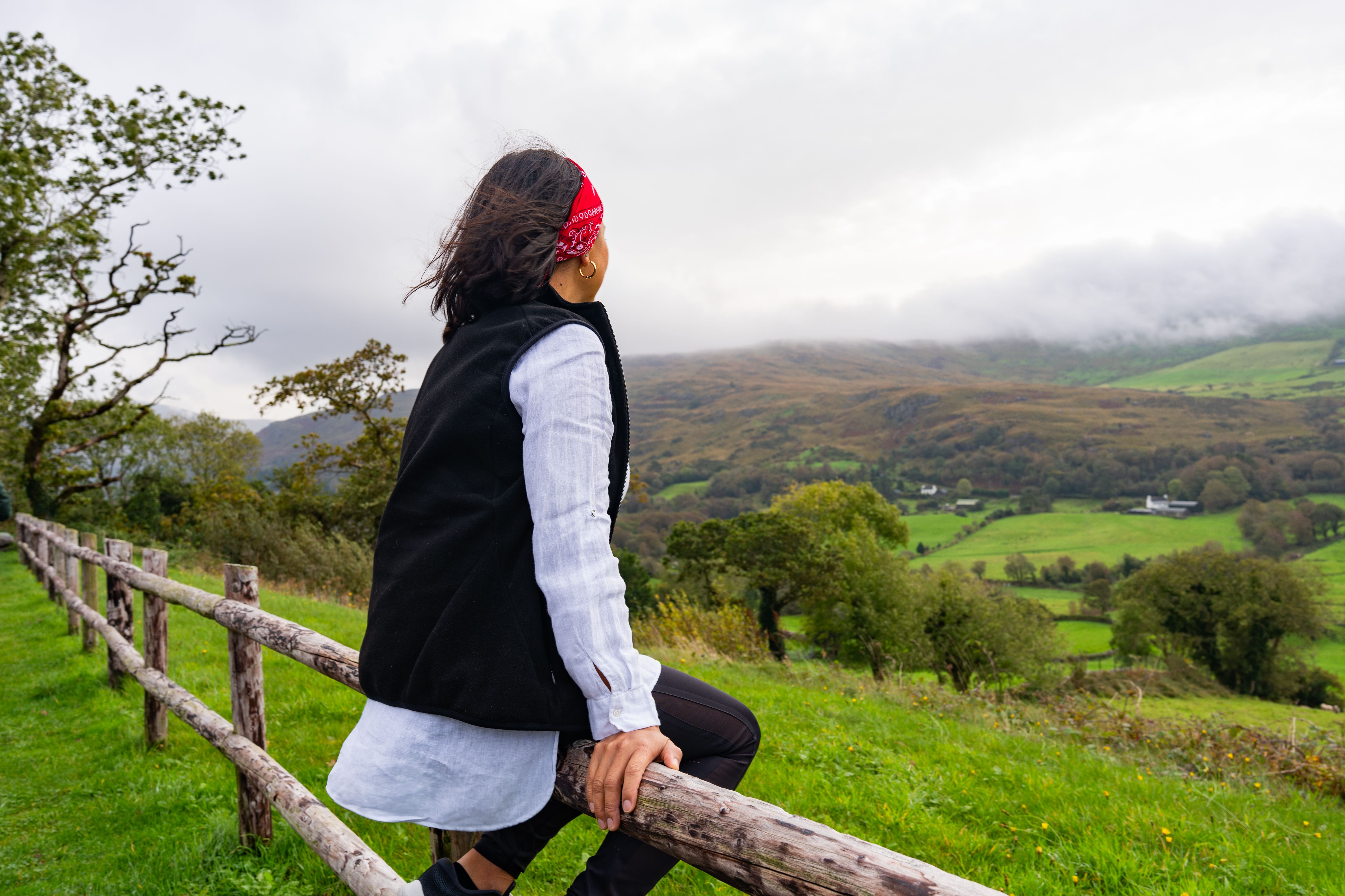 Young woman sitting on a wooden fence resting from their road trip through Ireland to enjoy a splendid landscape of green fields and low clouds