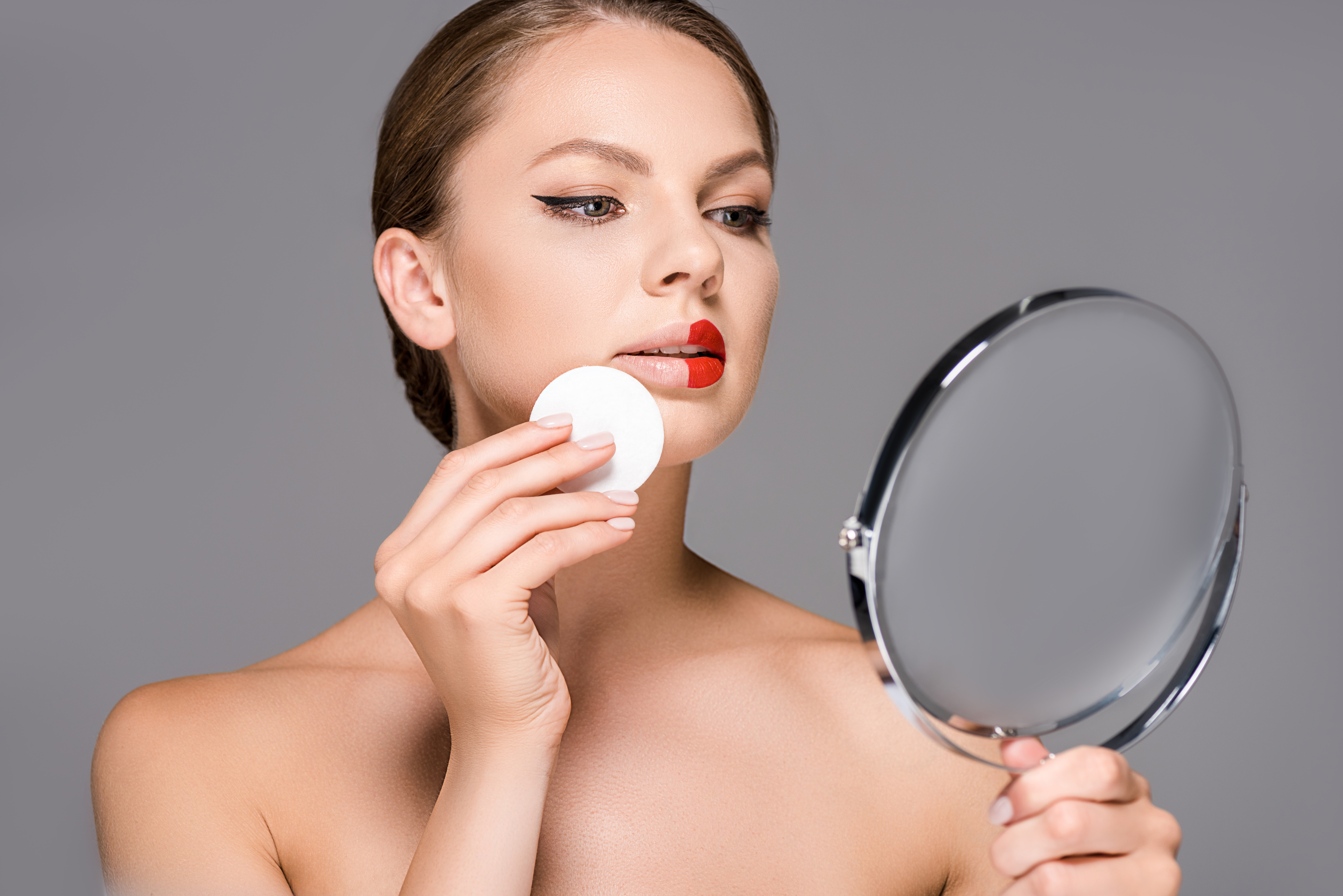 portrait of young woman with red lipstick on half of lips and sponge looking at mirror isolated on grey