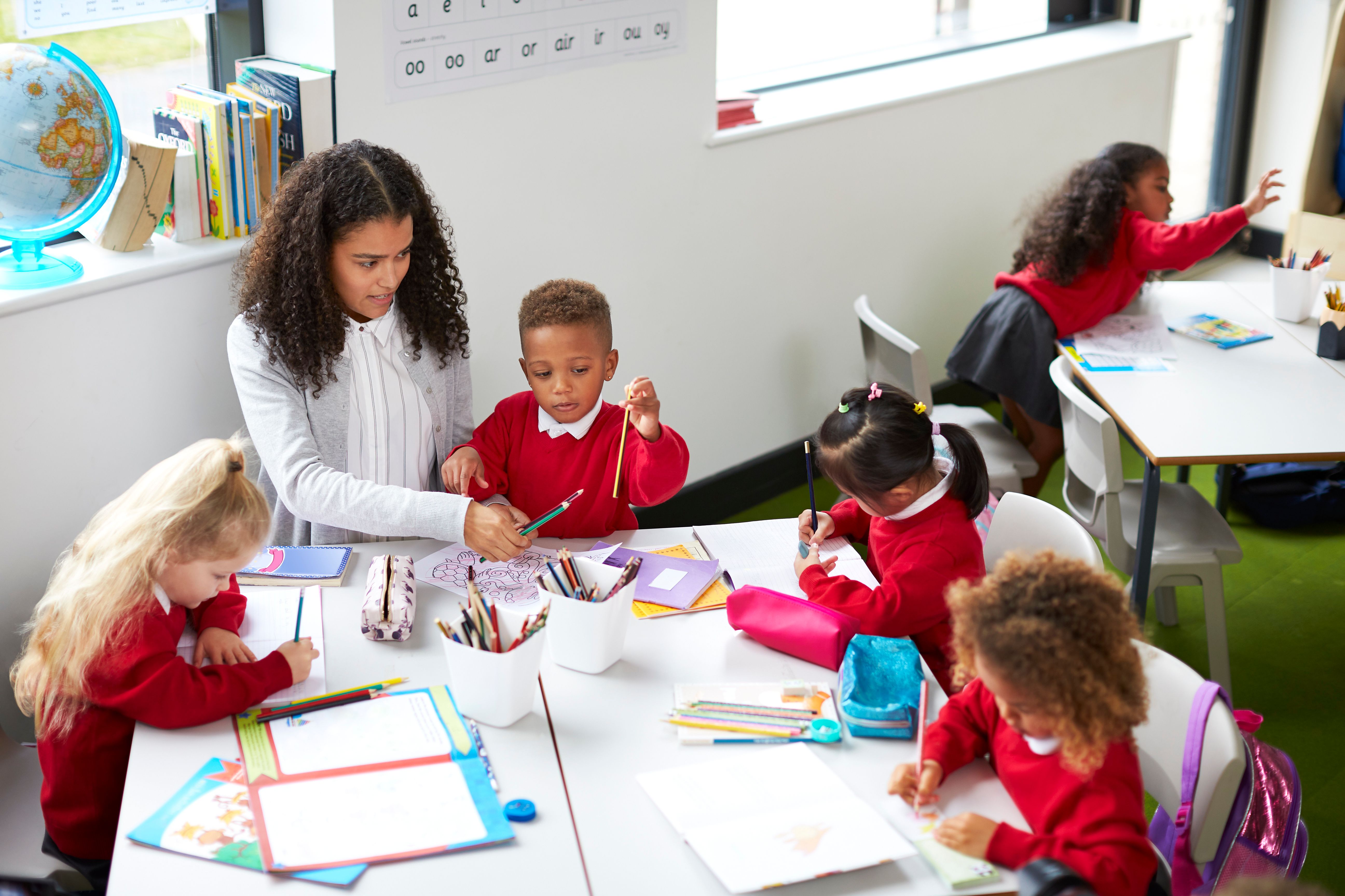 Elevated view of female kindergarten teacher sitting at a table helping four children during a lesson