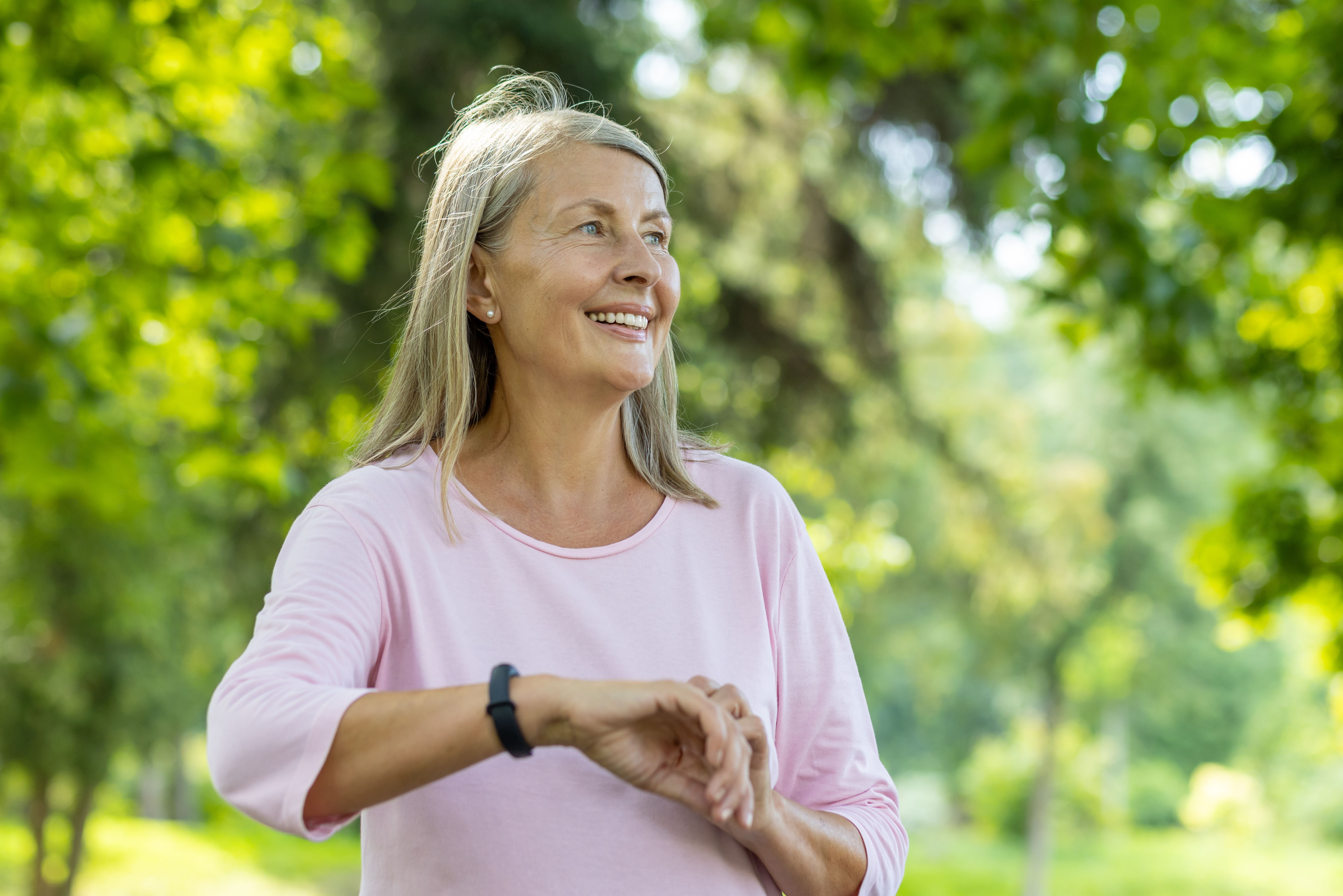 Senior beautiful gray-haired woman running in park, pensioner resting and using fitness bracelet of smart watch to choose sports programs, happy with active lifestyle near trees on sunny day