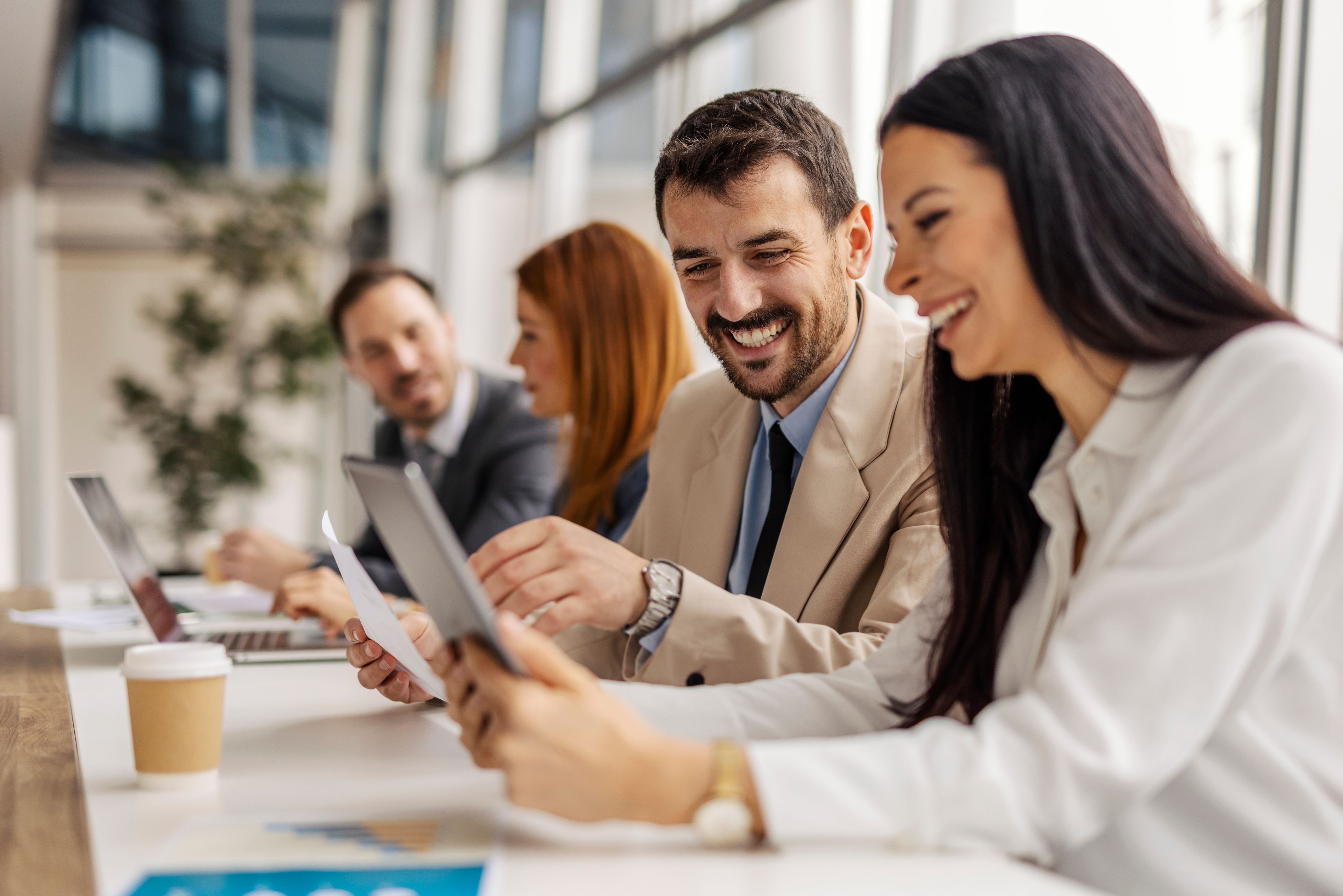 Two happy businesspeople smiling at tablet while sitting at boardroom on a meeting.