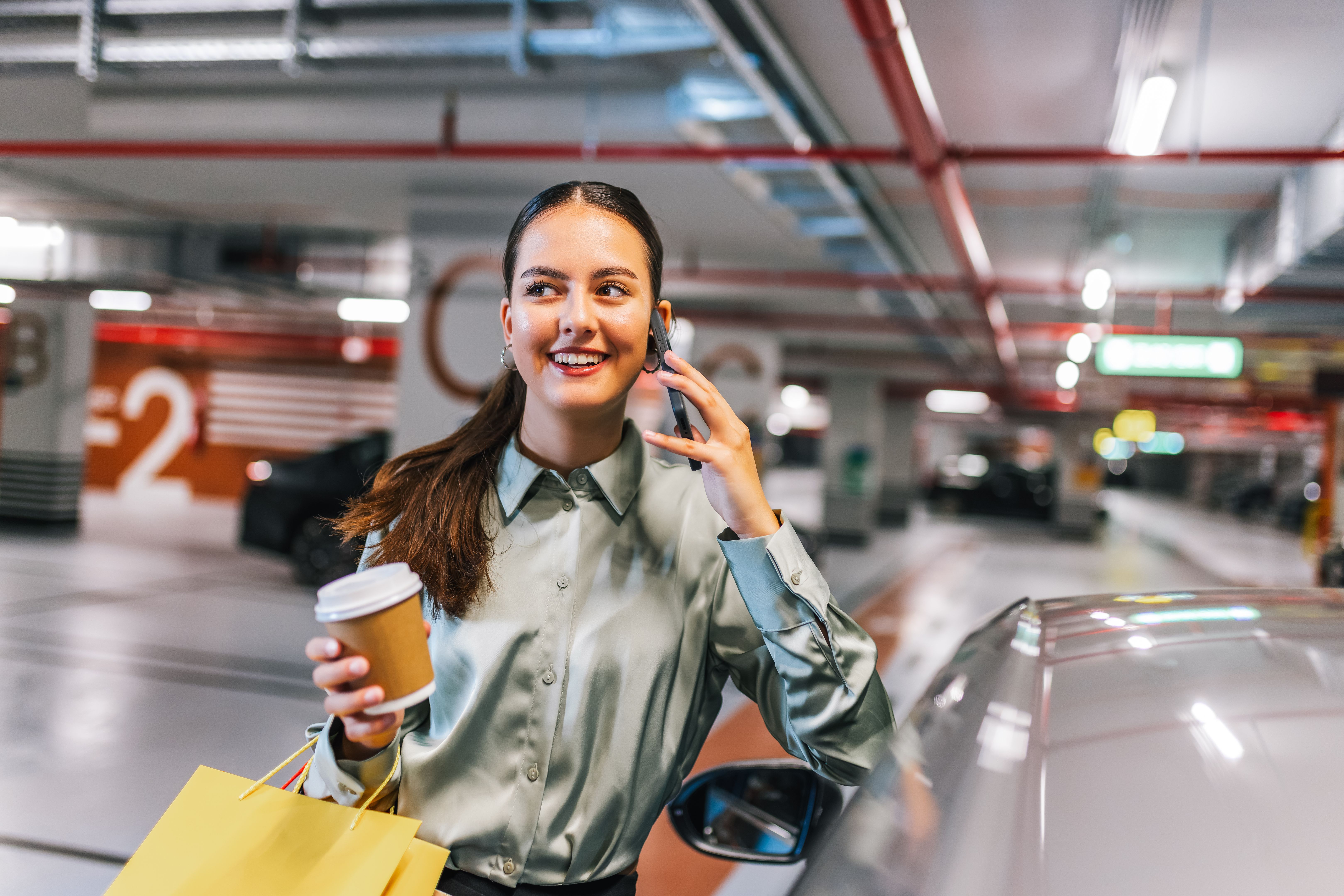 Young woman standing by her car with shopping bags using her phone and drinking coffee.