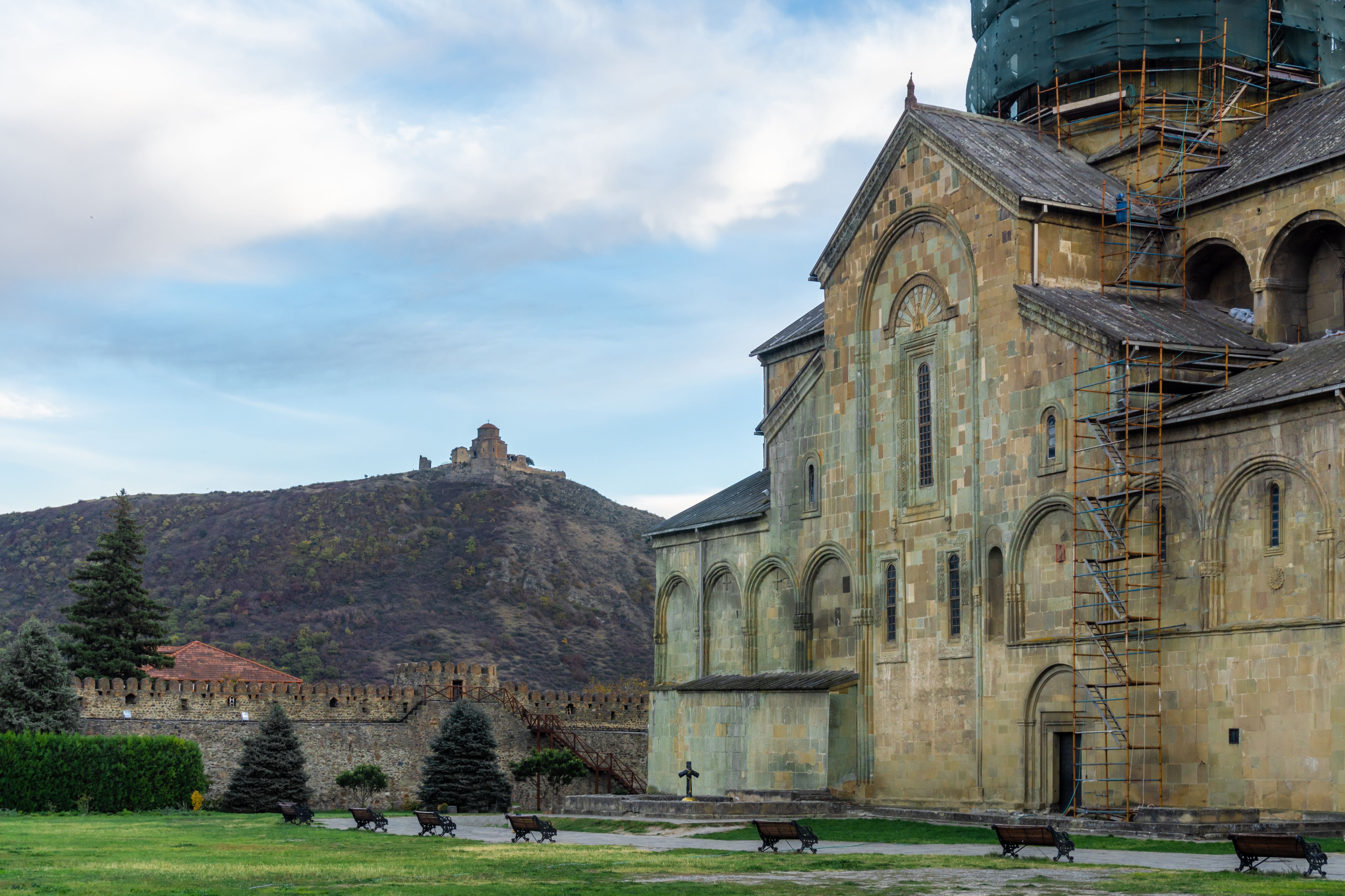 Svetitskhoveli Cathedral courtyard. Left wall of the church, green lawn, benches, seats. Jvari Monastery on the hill in the background. Bright sky