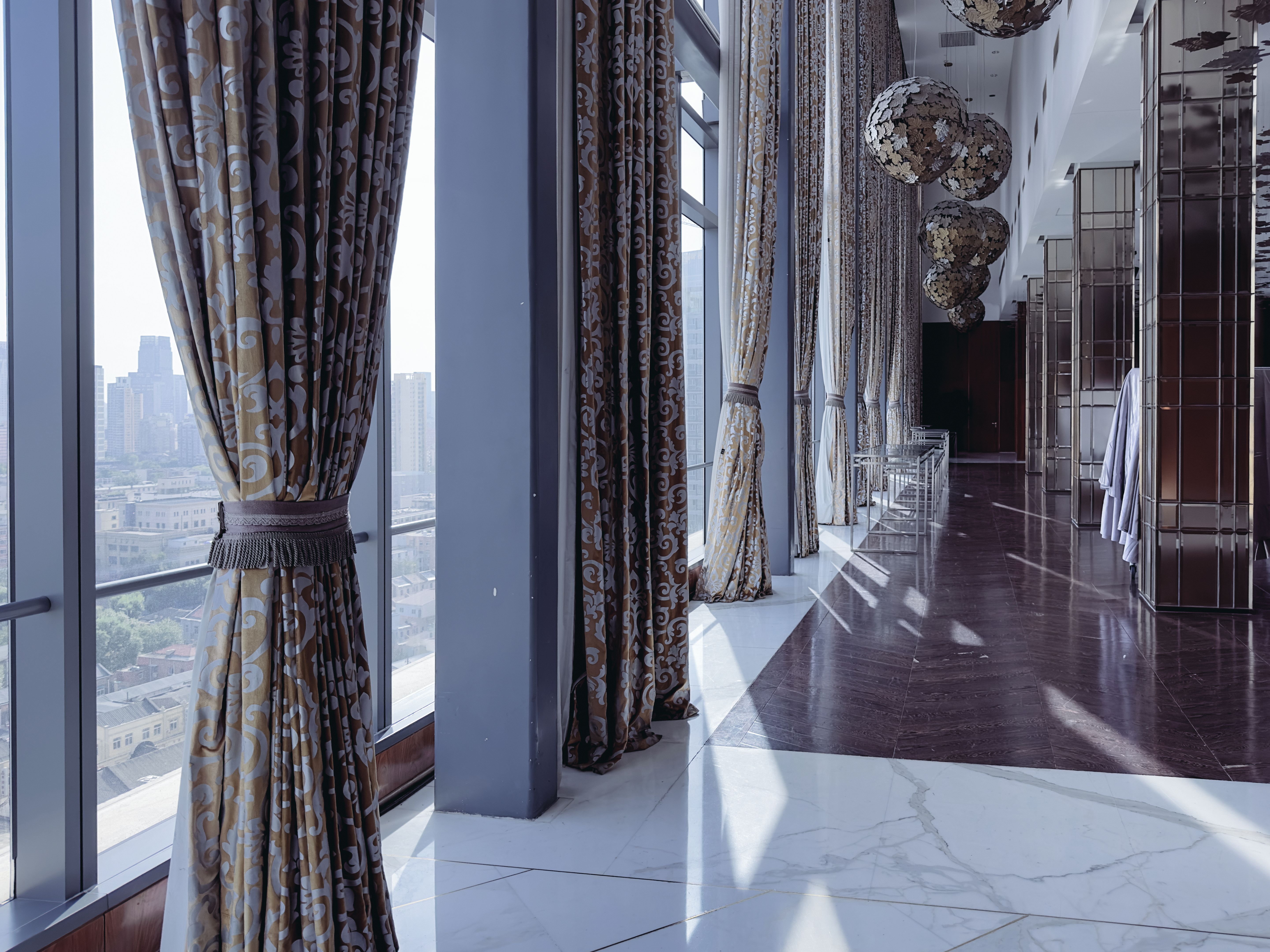 A spacious, modern interior of a high-rise building’s event or conference room. The space features large floor-to-ceiling windows with flowing, elegant curtains, allowing natural light to flood the room.