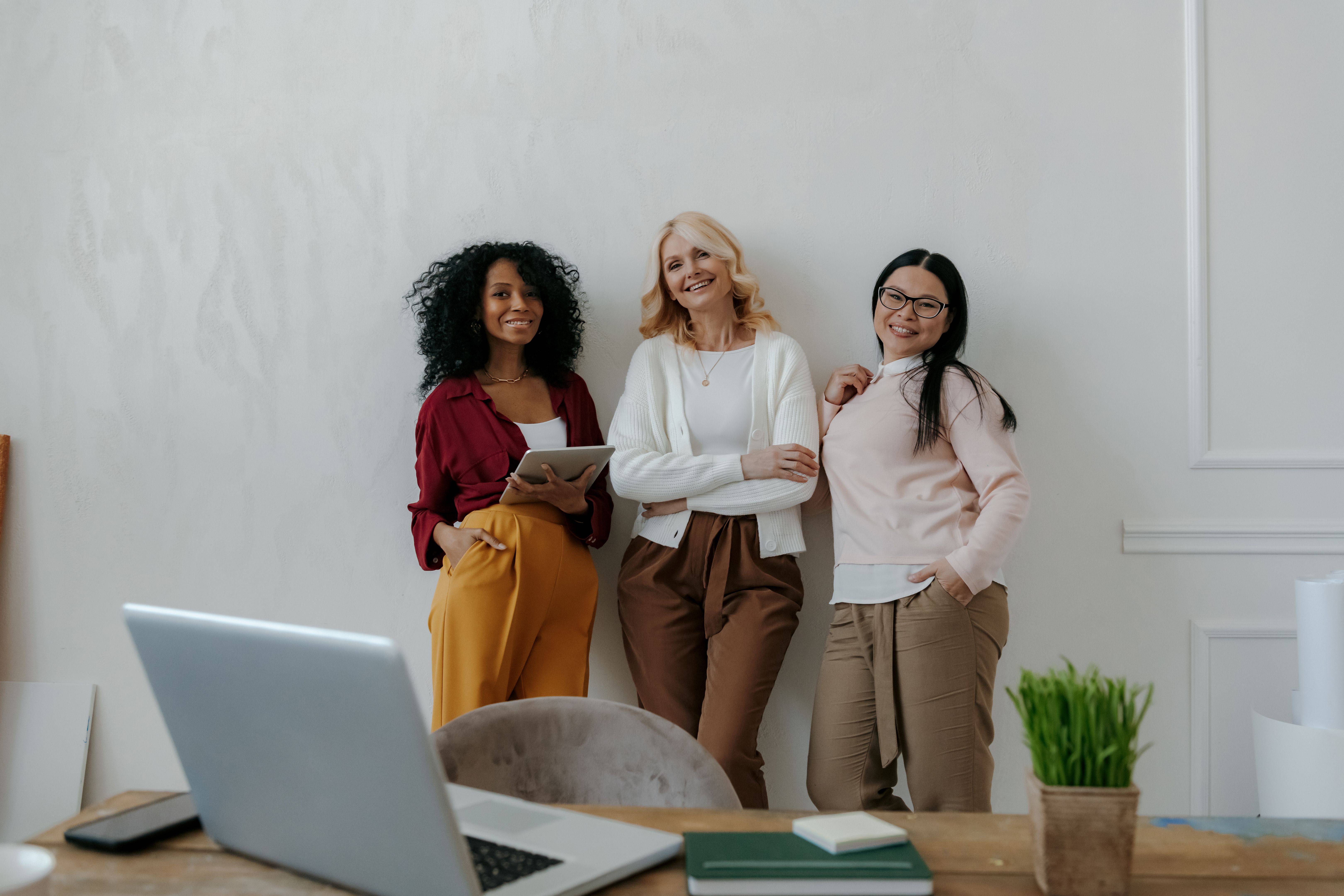 Three modern mature women smiling while leaning on the wall in office