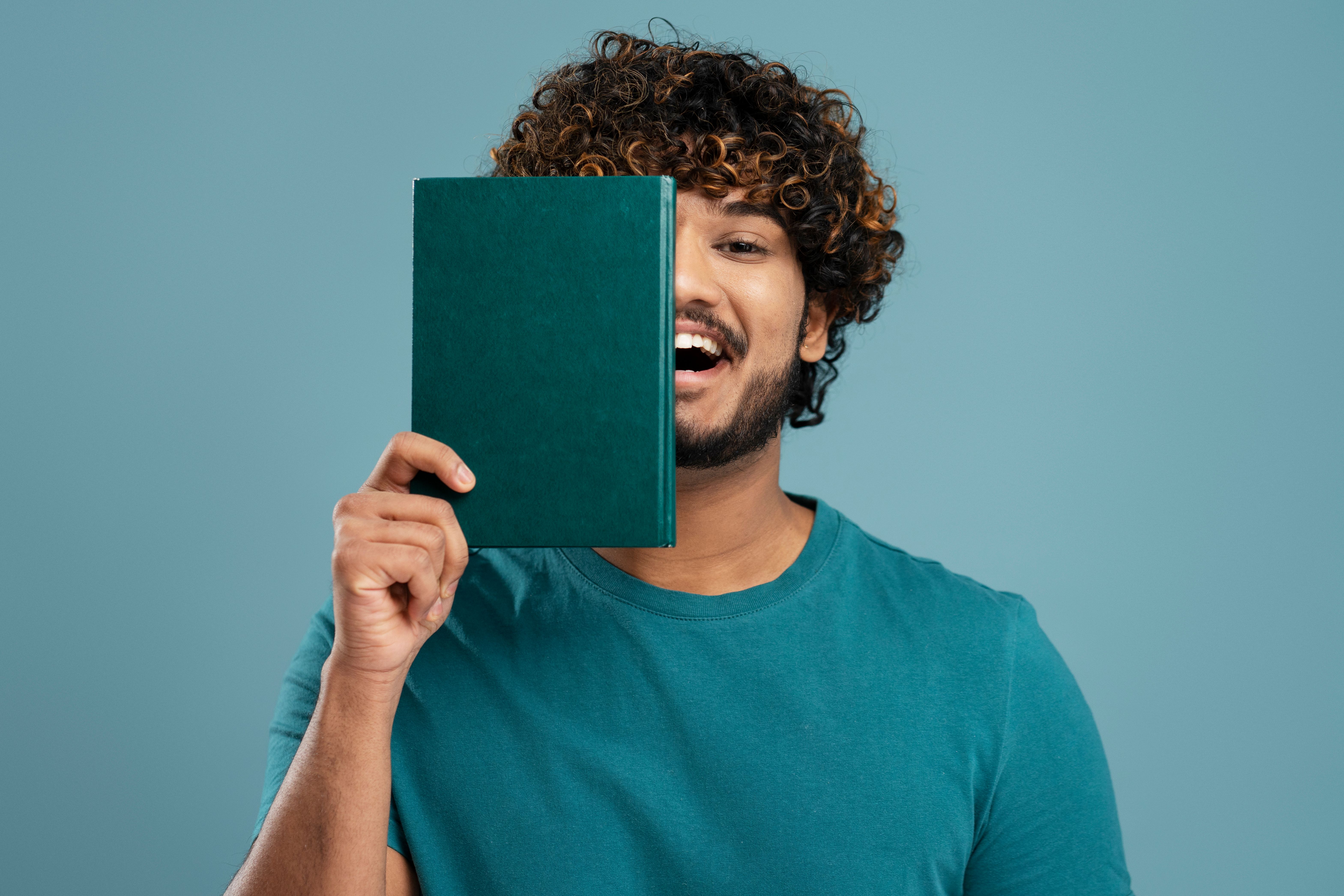 Cheerful Indian male student smiling and covering half of his face with a green hardcover book