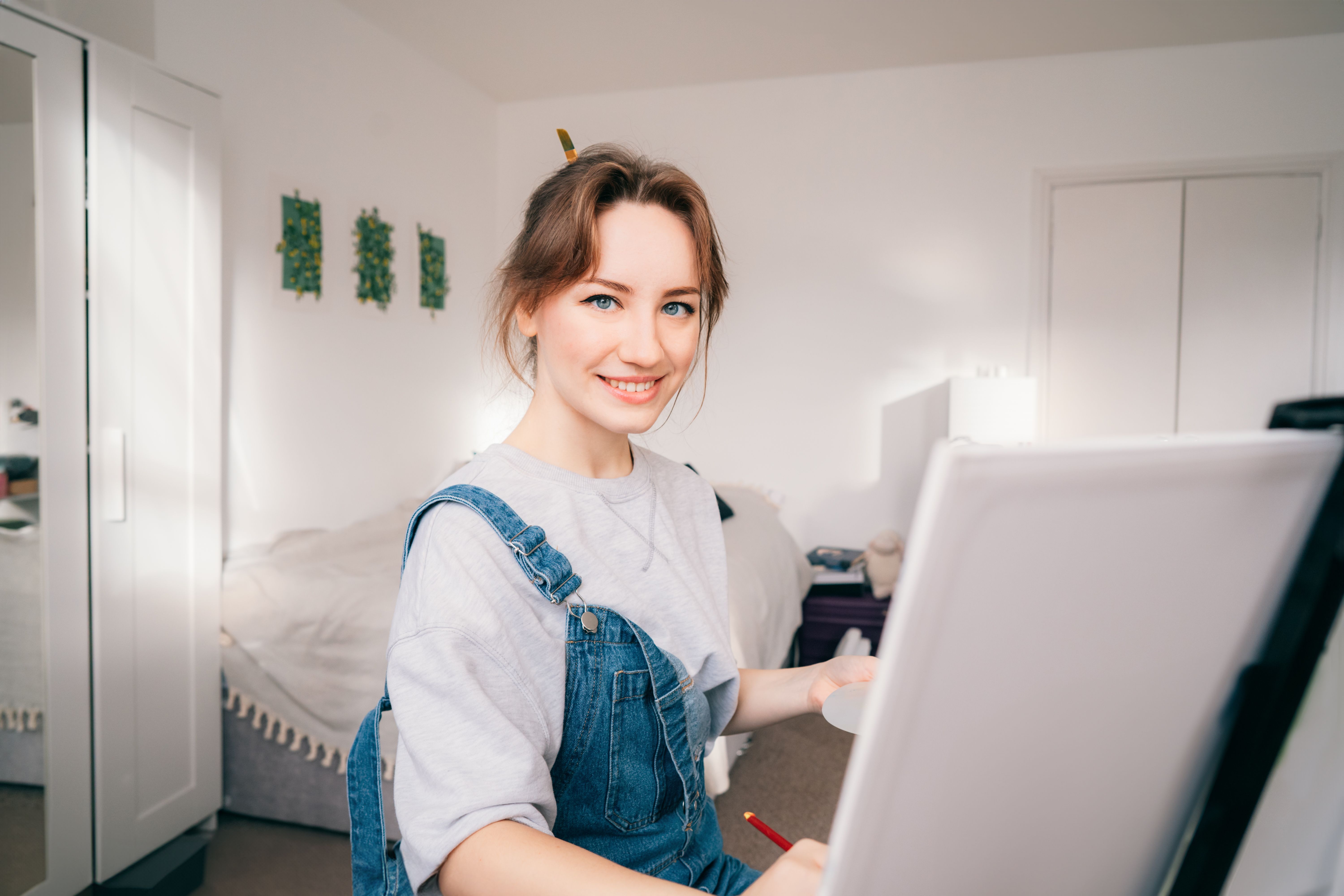 female artist sitting in front of easel, painting picture with inspiration, drawing on canvas with paintbrush, palette, smiling, relaxing at home. Art, hobby, leisure and creative occupation concept