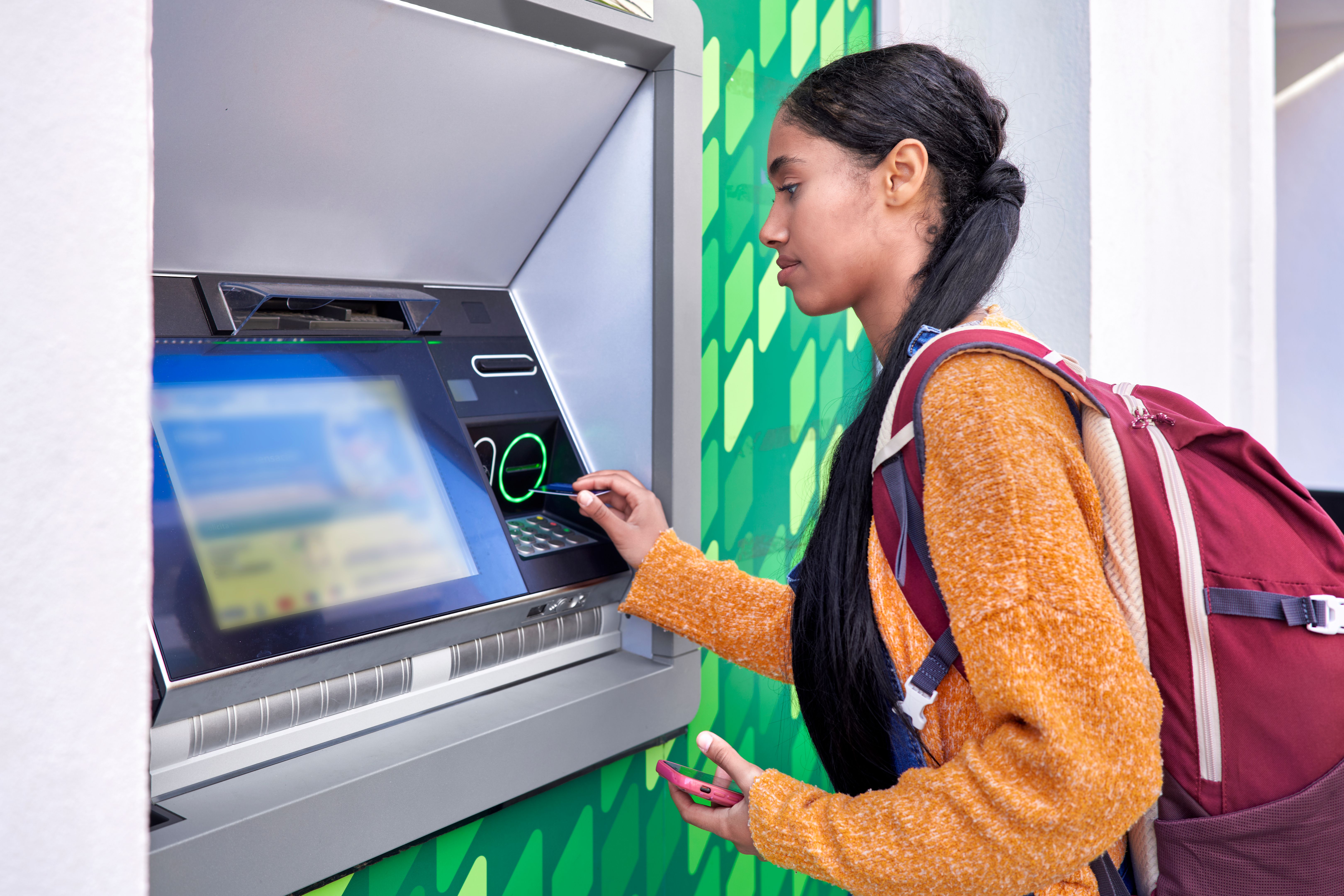Young and pretty digital nomad Colombian Latin girl with her backpack scanning direro with her credit card at an ATM Young and pretty digital nomad Colombian Latin girl with her backpack scanning direro with her credit card at an ATM