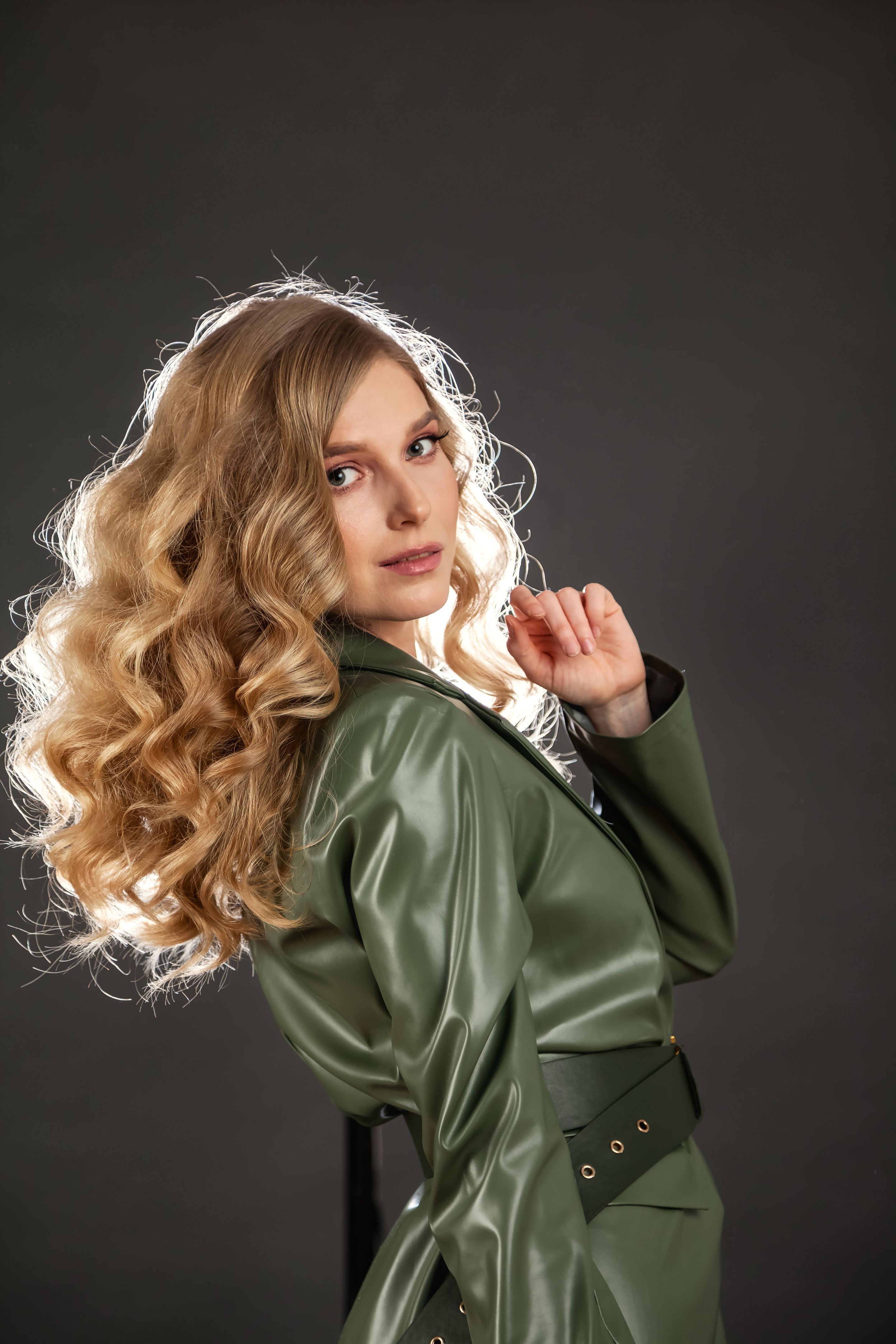 Cute young woman in green jacket posing at dark grey background, looking at camera. Studio shot of caucasian blonde stylish woman with curly hair. Dance psychology concept. Copy text space for ad
