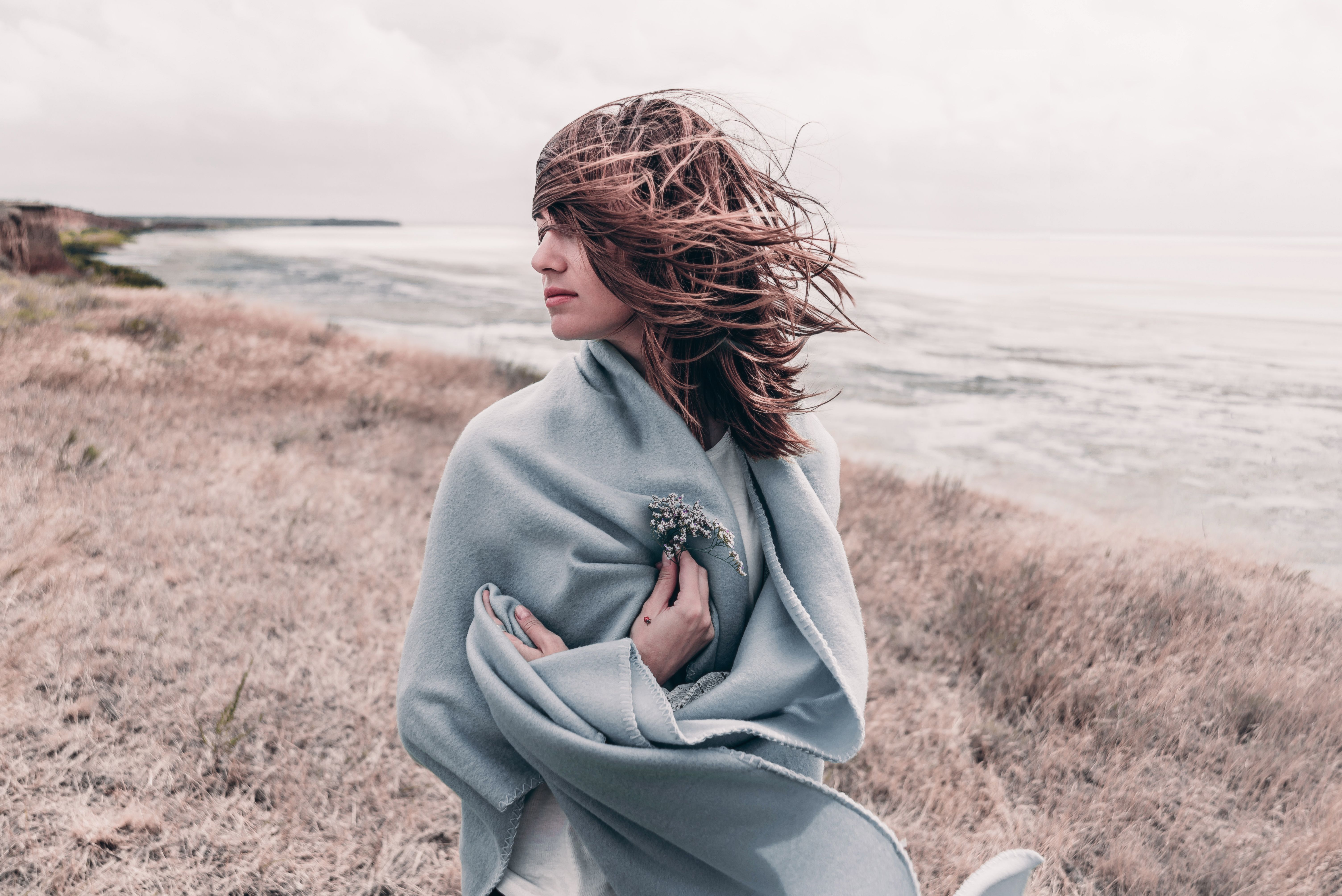 Attractive young woman standing on a windy cold beach wrapped in warm blanket with flower in her hand. Attractive young woman standing on a windy cold beach wrapped in warm blanket with flower in her hand.