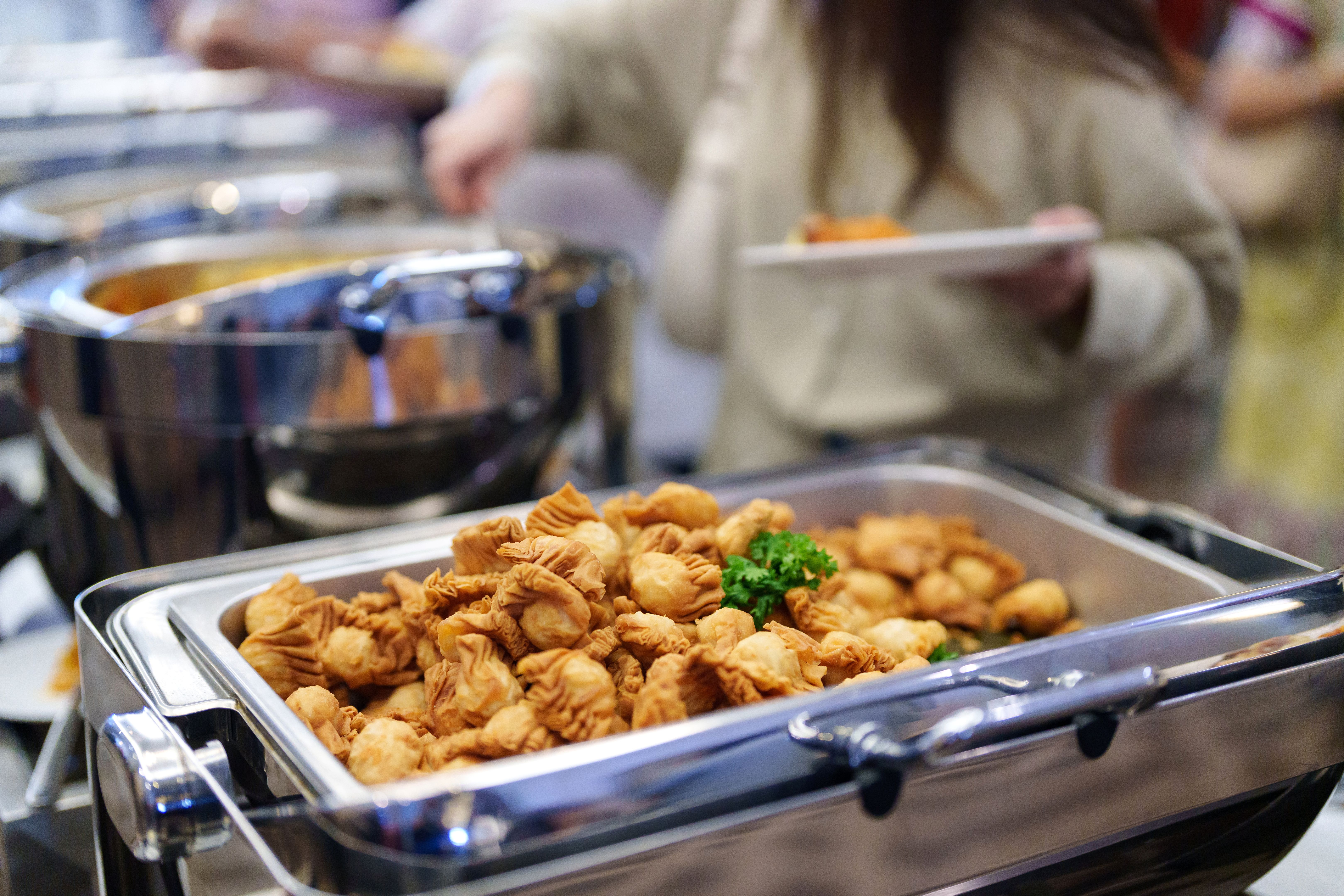 A cropped image captures businesspeople in line, using serving spoons to pick their food from the buffet. A cropped image captures businesspeople in line, using serving spoons to pick their food from the buffet.