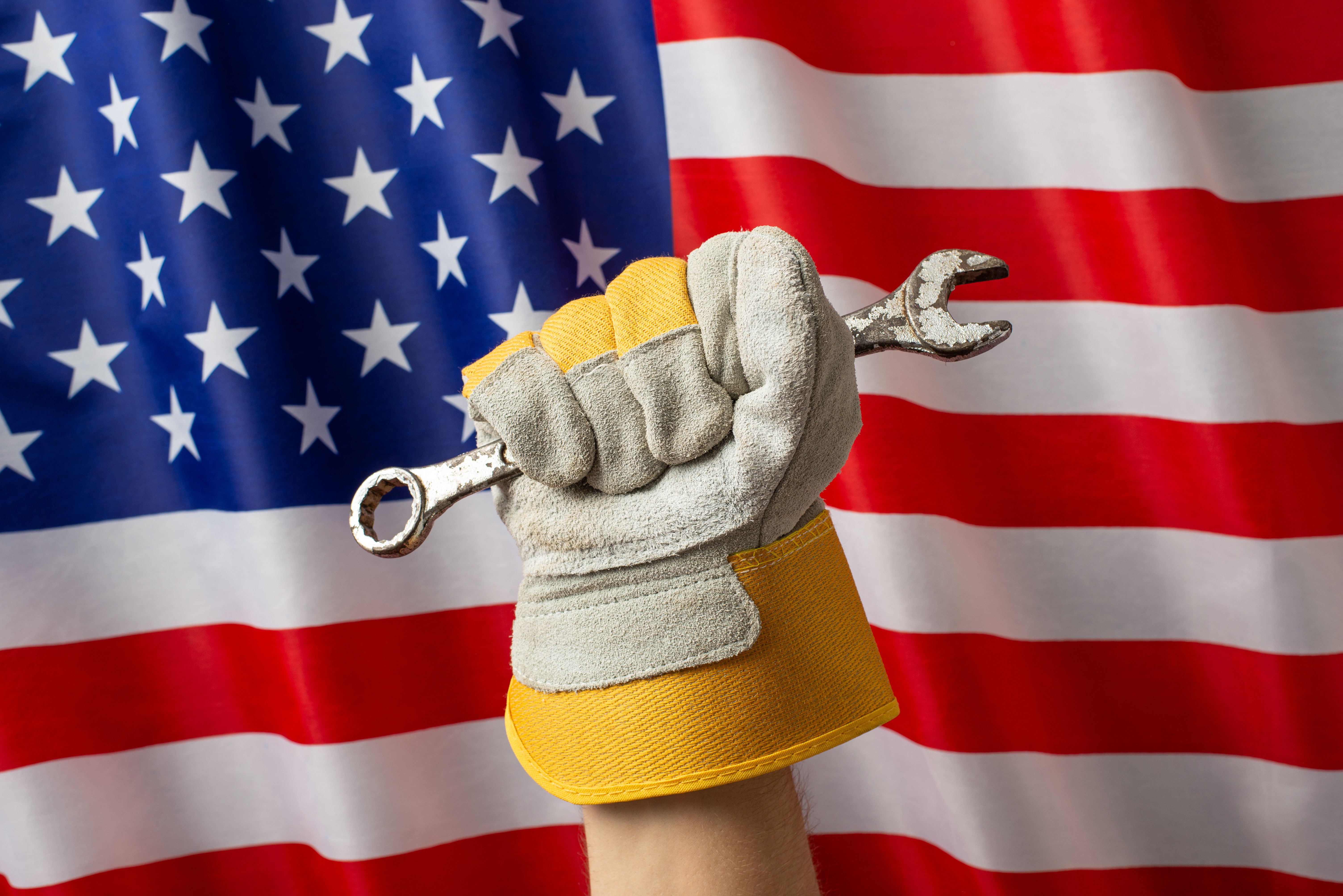 A gloved hand holding a wrench in front of an American flag symbolizes labor, strength, and patriotism