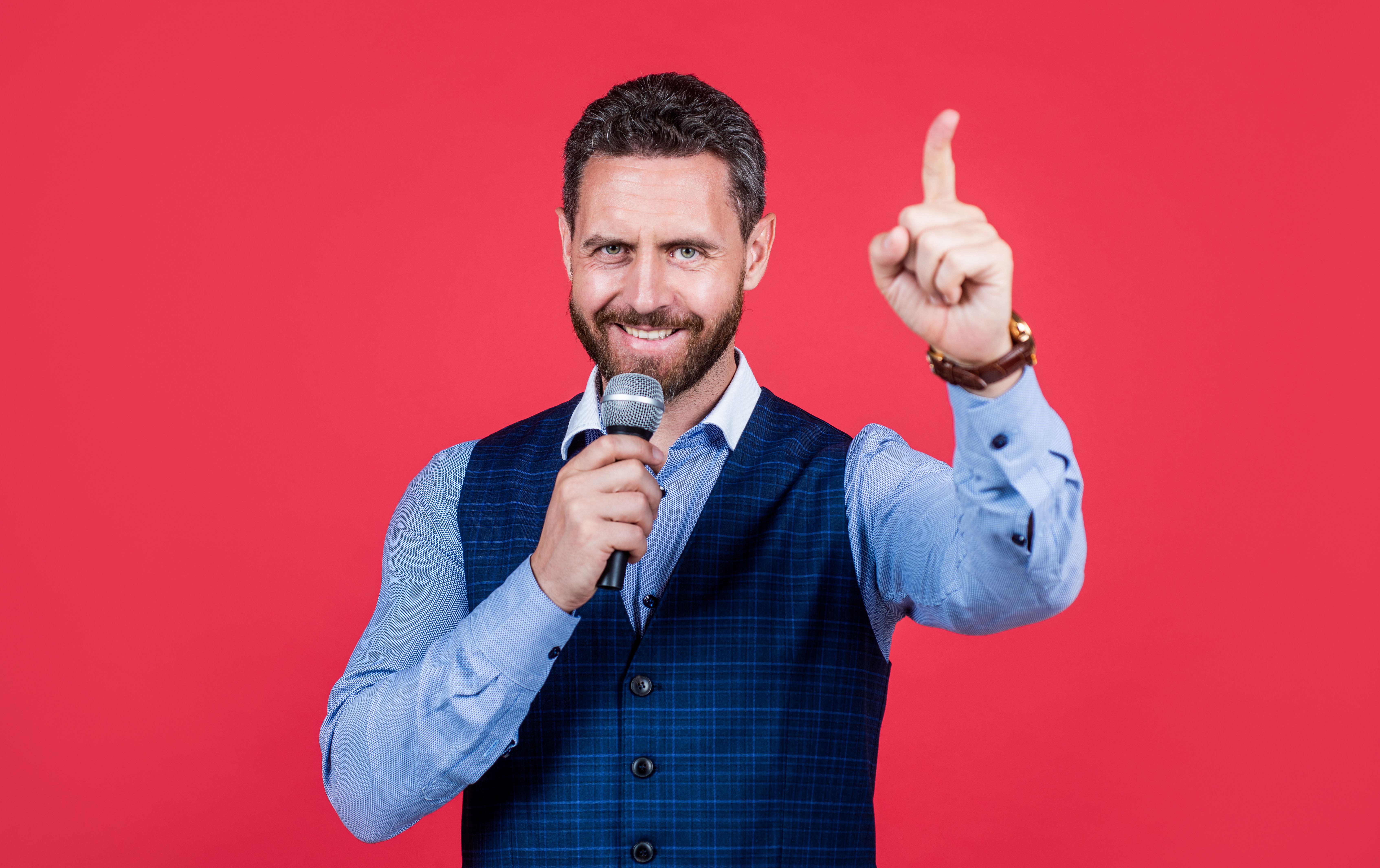 Talk show host. Happy speaker speak into microphone keeping finger raised. Speaker perform speech. Visiting professor or lecturer. Speaking coach. Hosting conference. Voice professional