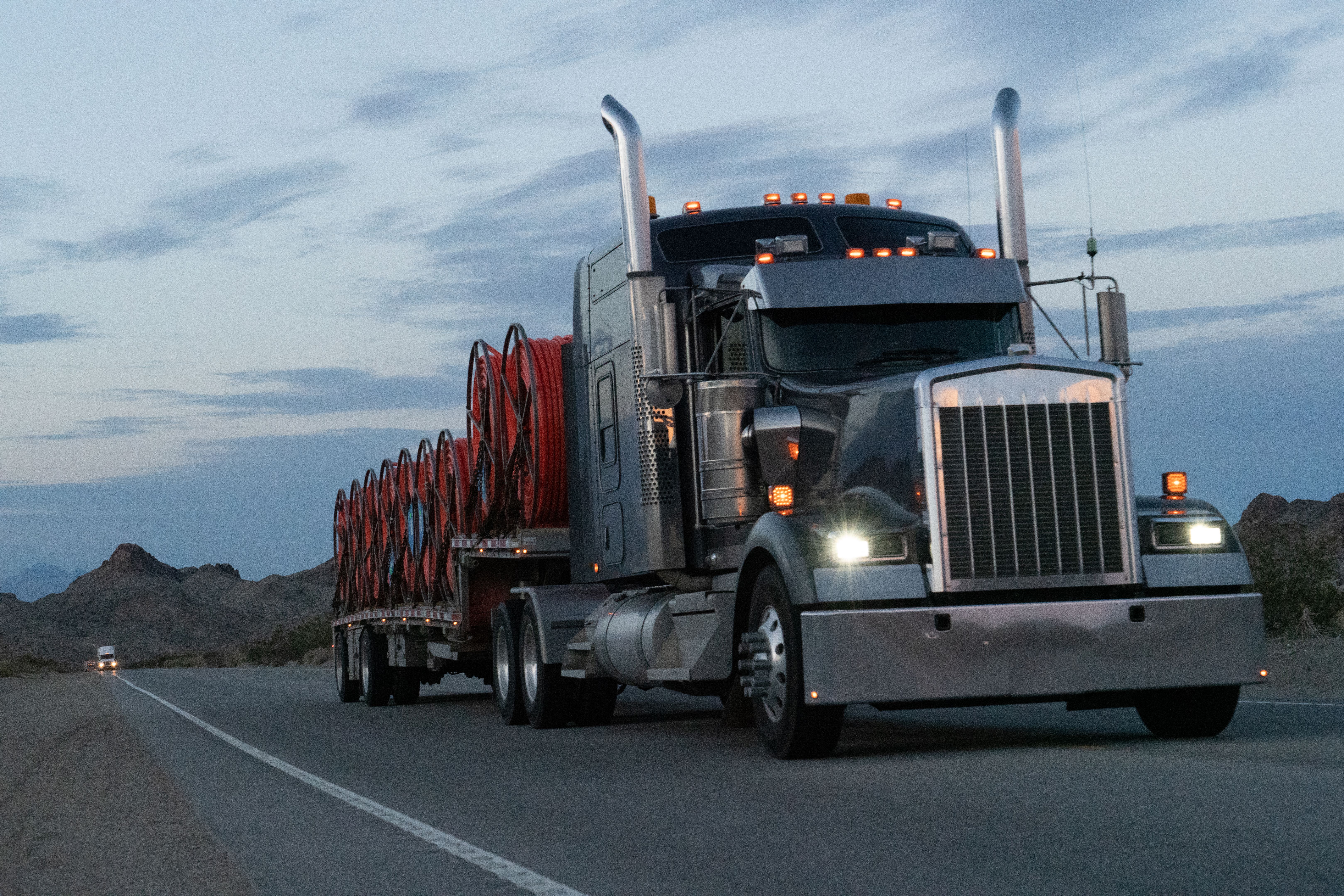 As the sun dipped below the horizon, a semi truck rumbled through the Nevada desert. Its powerful silhouette cut through the vast landscape, with majestic mountains standing as a breathtaking backdrop.