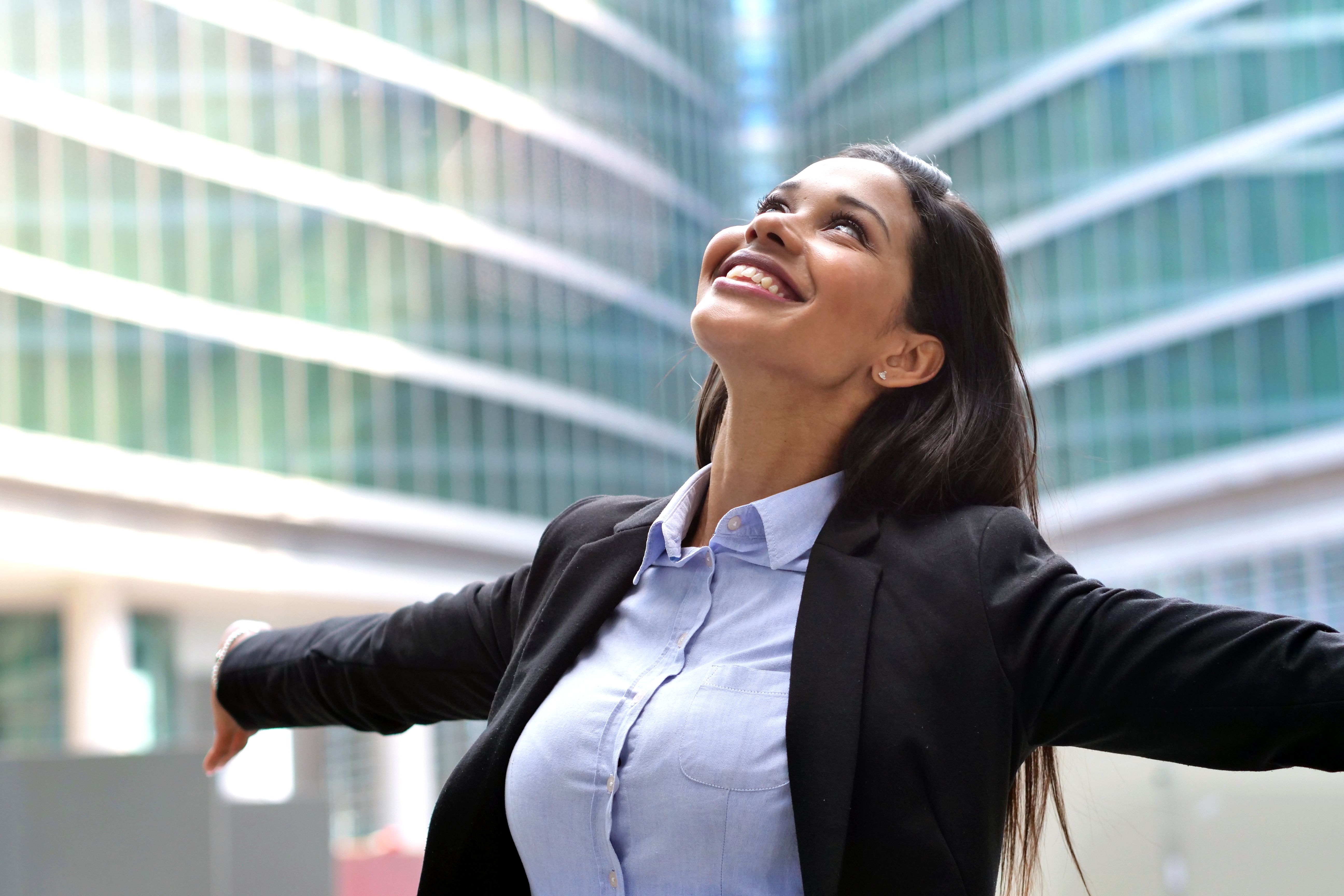 Una mujer africana de negocios está levantando los brazos hacia el cielo y respira en un fondo de rascacielos como señal de libertad y éxito en su trabajo.