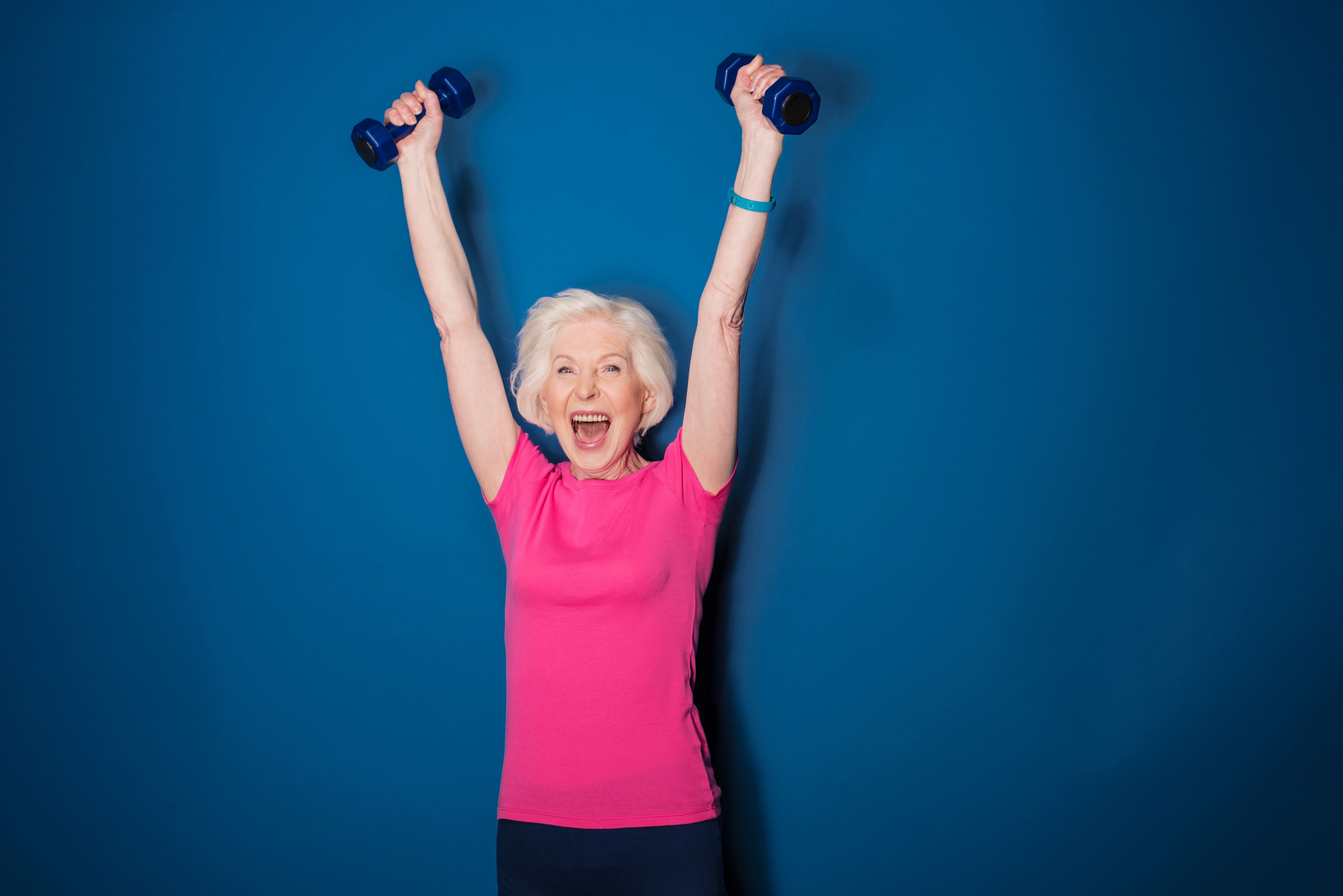 Excited senior fitness woman training with dumbbells isolated on blue