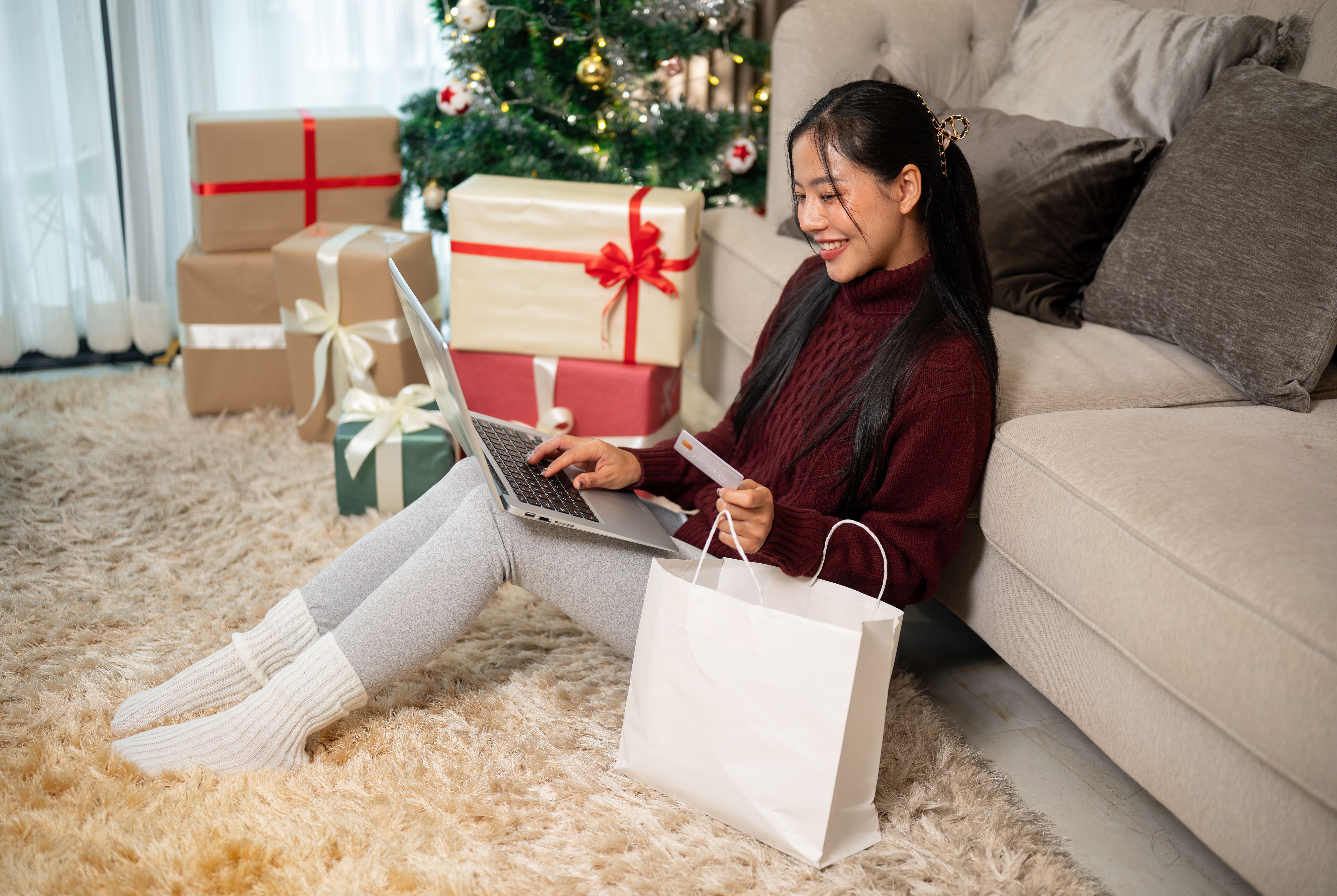 A happy Asian woman in cozy clothes enjoys shopping online at home during the Christmas holiday.