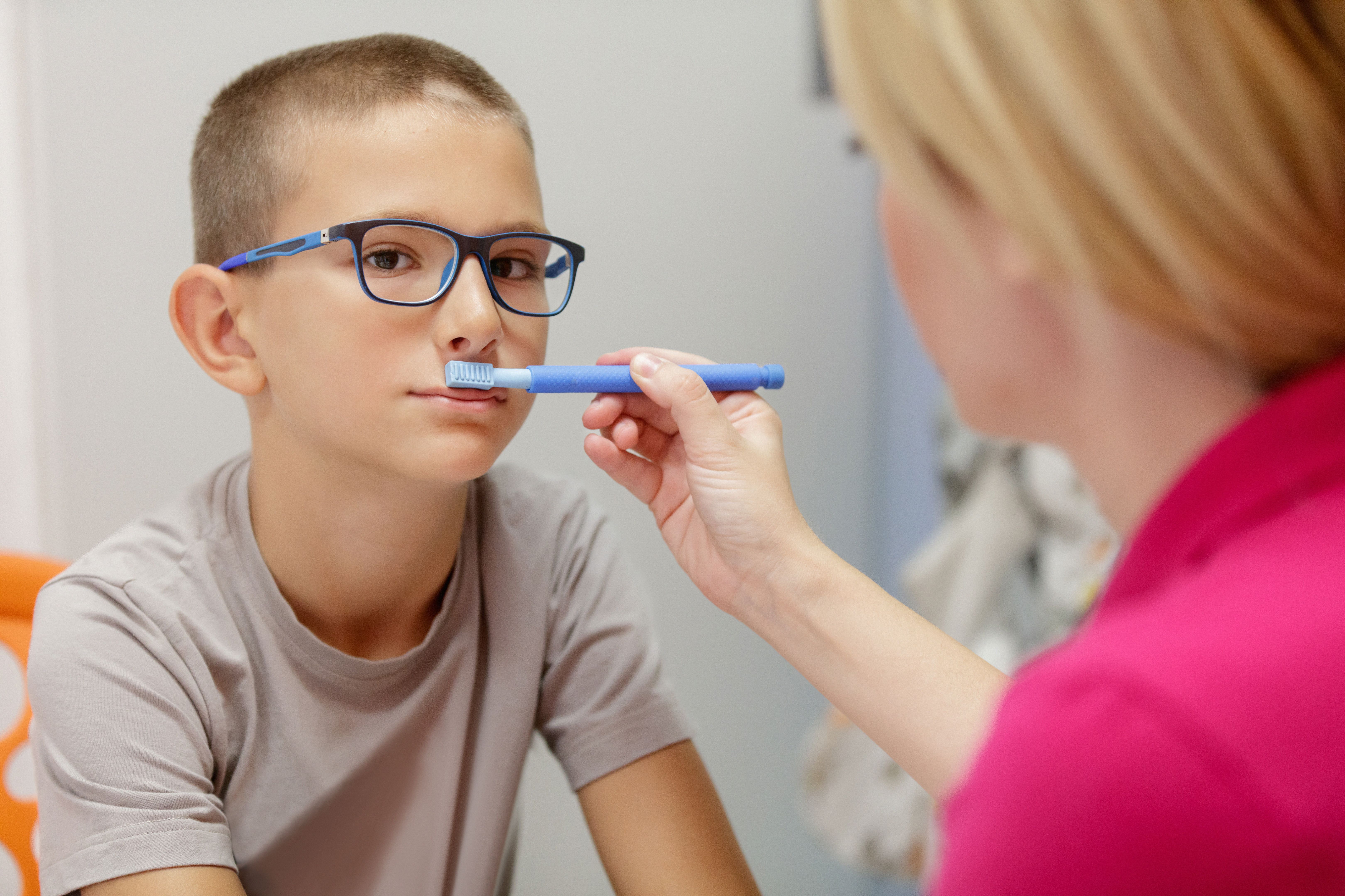 Female doctor using special aid in occupational therapy and massaging elementary age boy's upper lip muscles