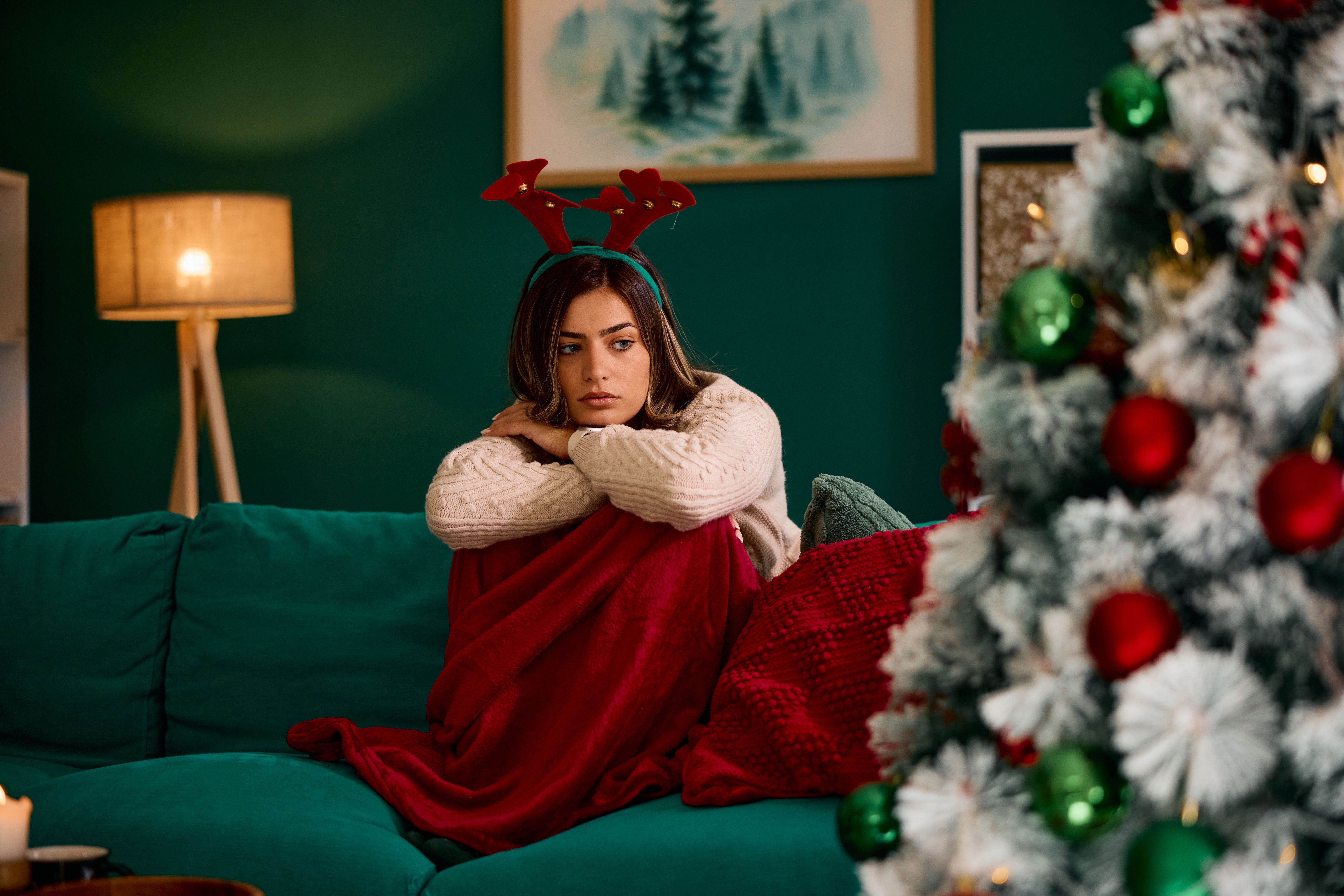 Sad young woman wearing reindeer antlers is sitting alone by the Christmas tree