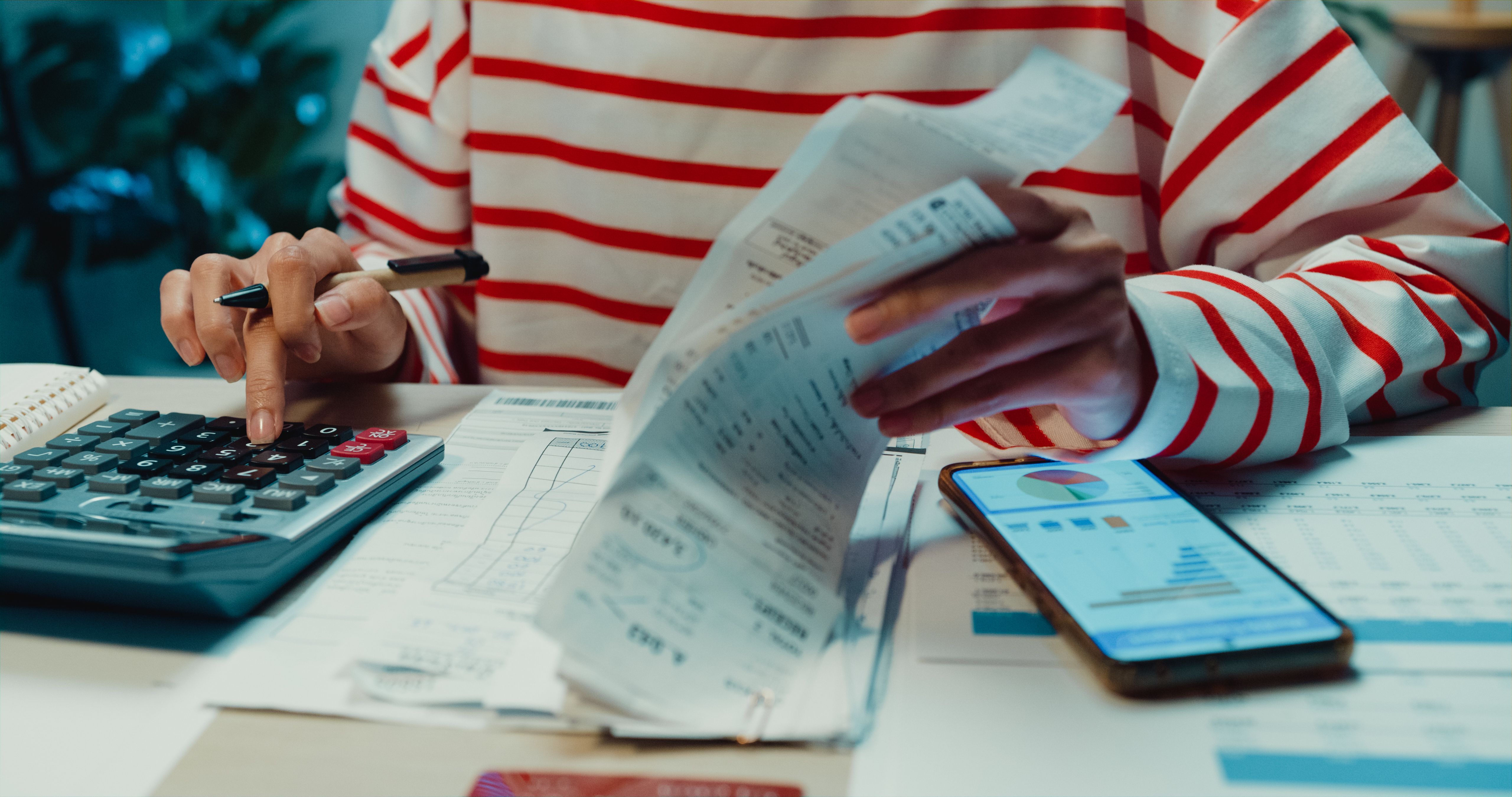 Close-up Young Asia woman with sweater sit in front of desk with phone use calculator to calculate utility bills check credit card receipt monthly expense bill in house at night. Close-up Young Asia woman with sweater sit in front of desk with phone use calculator to calculate utility bills check credit card receipt monthly expense bill in house at night.