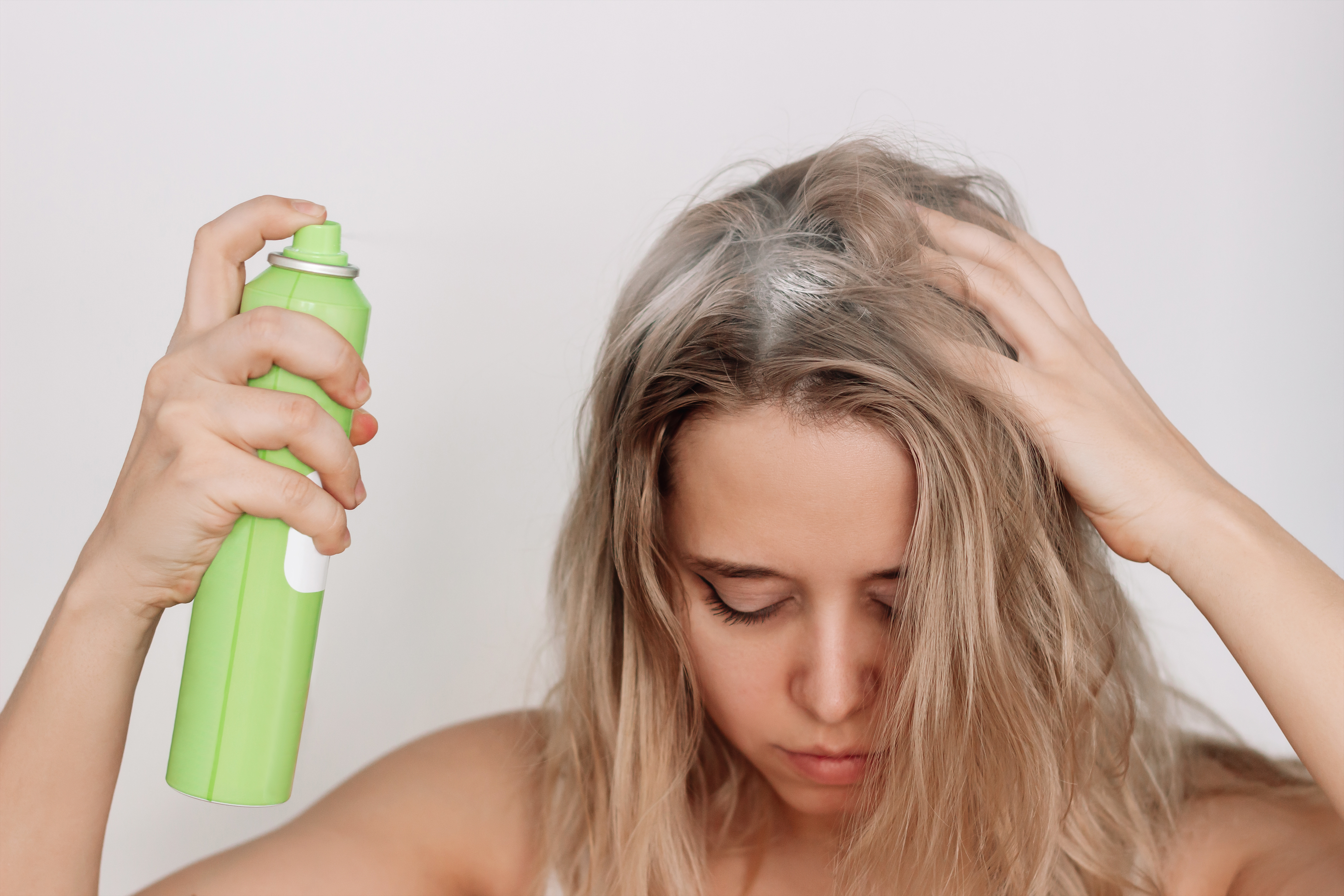 Young blonde woman with dirty greasy hair spraying a dry shampoo on the roots of her hair