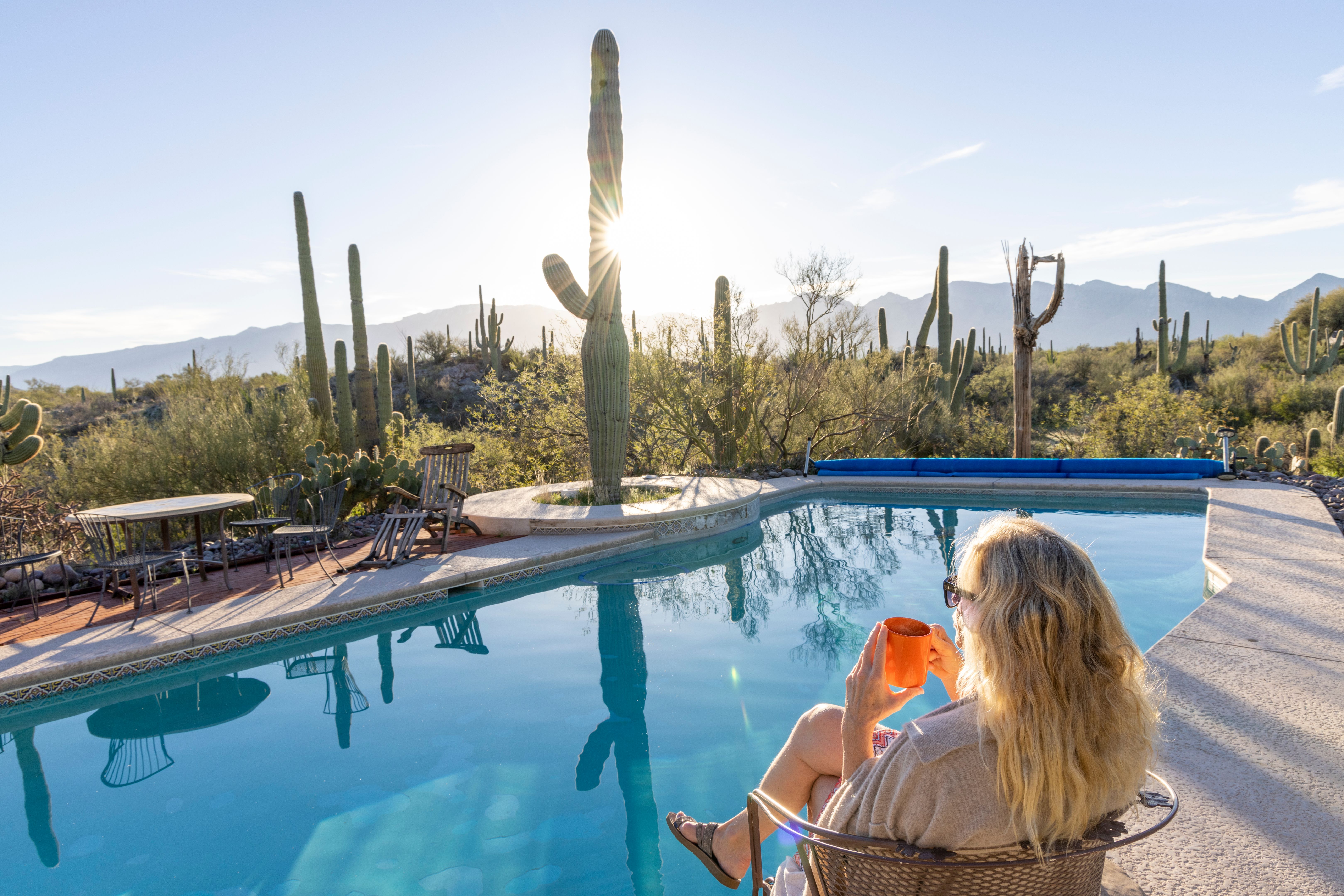 Blonde woman relaxes with coffee by swimming pool looking out across an open field of saguaros and lush Sonora Desert landscape after a Monsoon Blonde woman relaxes with coffee by swimming pool looking out across an open field of saguaros and lush Sonora Desert landscape after a Monsoon