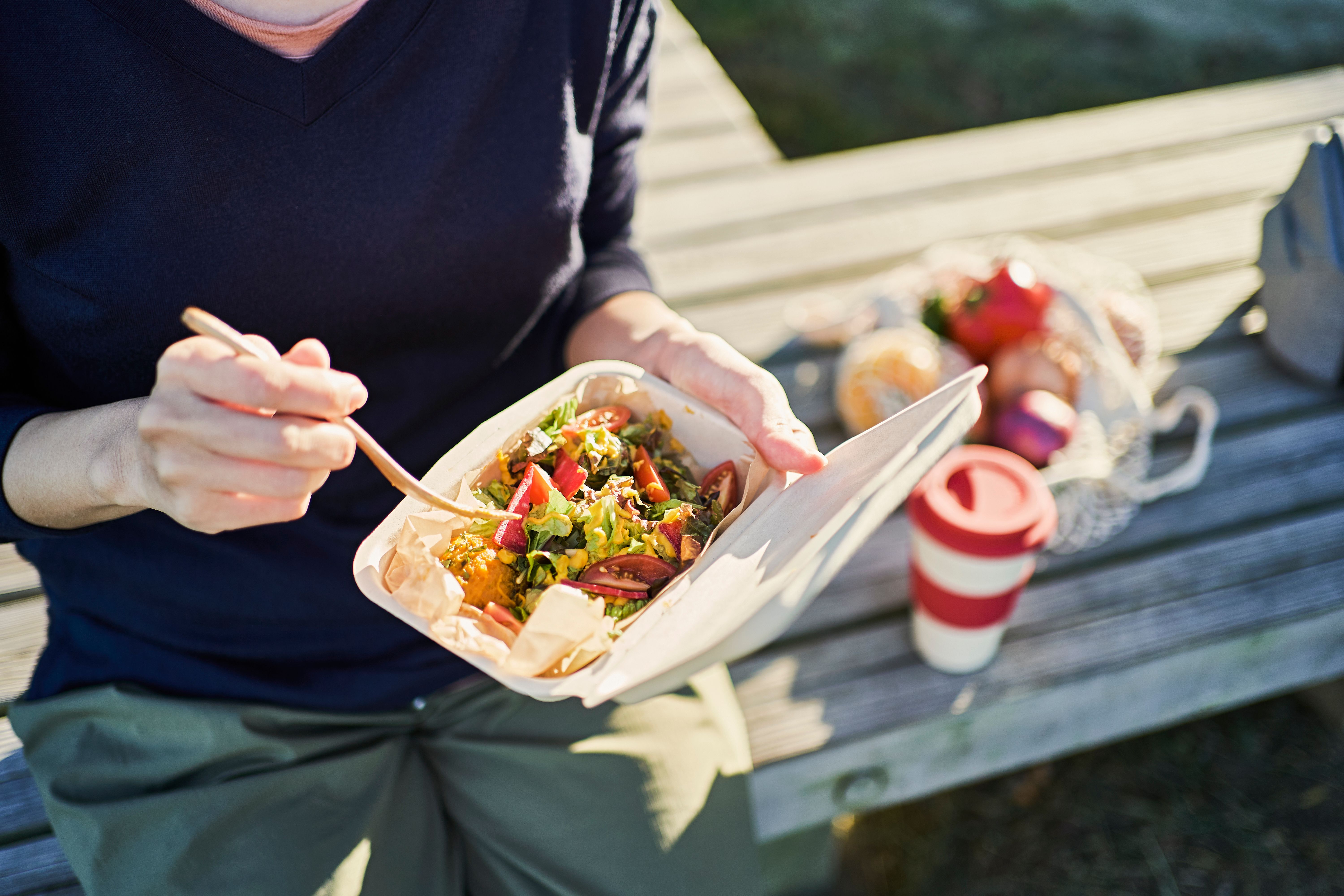 A woman is outside on a bench eating from a compostable to go sugarcane hinged box with a sunny background A woman is outside on a bench eating from a compostable to go sugarcane hinged box with a sunny background