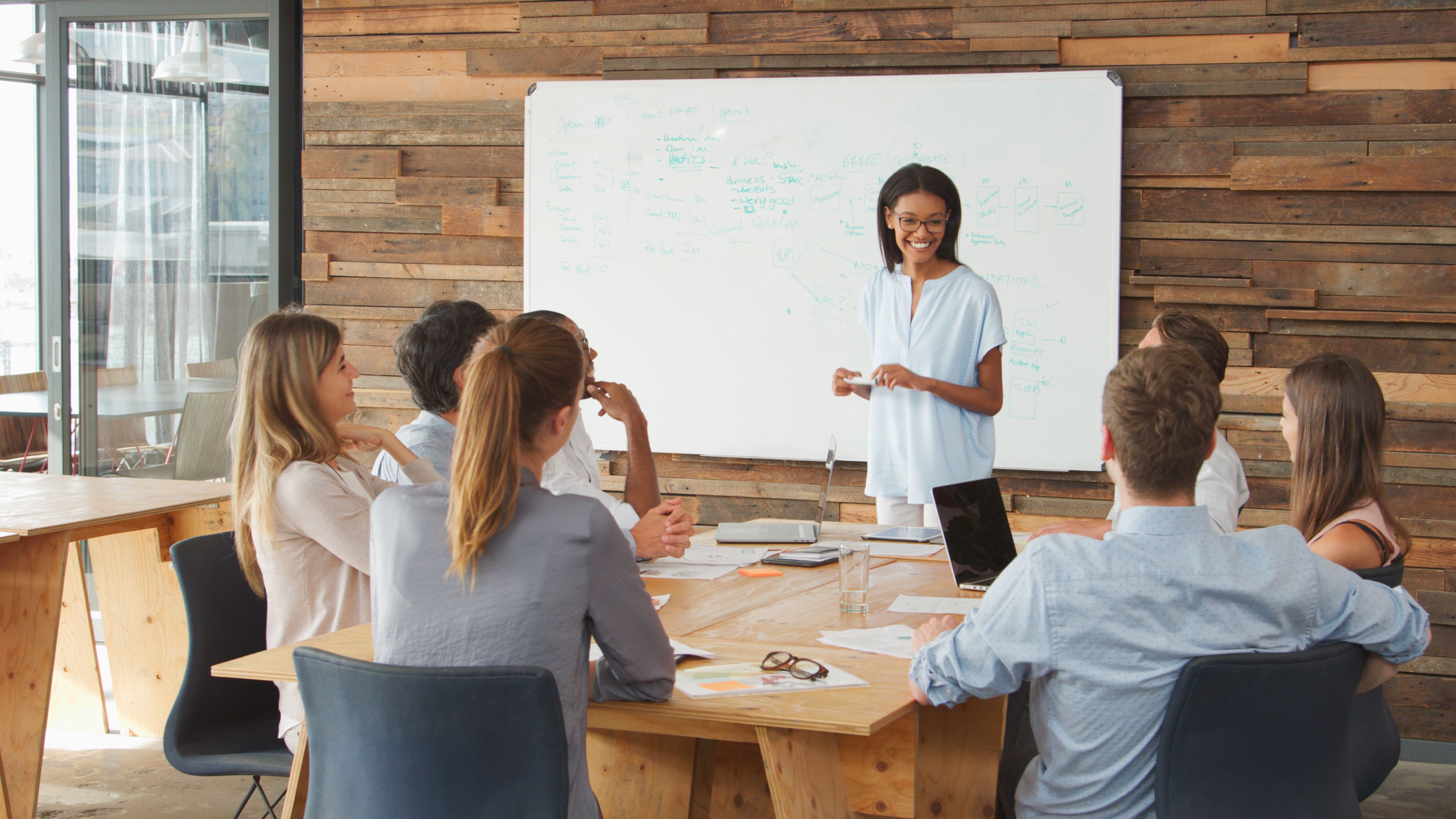 Young Businesswoman In Front Of Whiteboard Making Presentation To Colleagues Around Table In Office Young Businesswoman In Front Of Whiteboard Making Presentation To Colleagues Around Table In Office