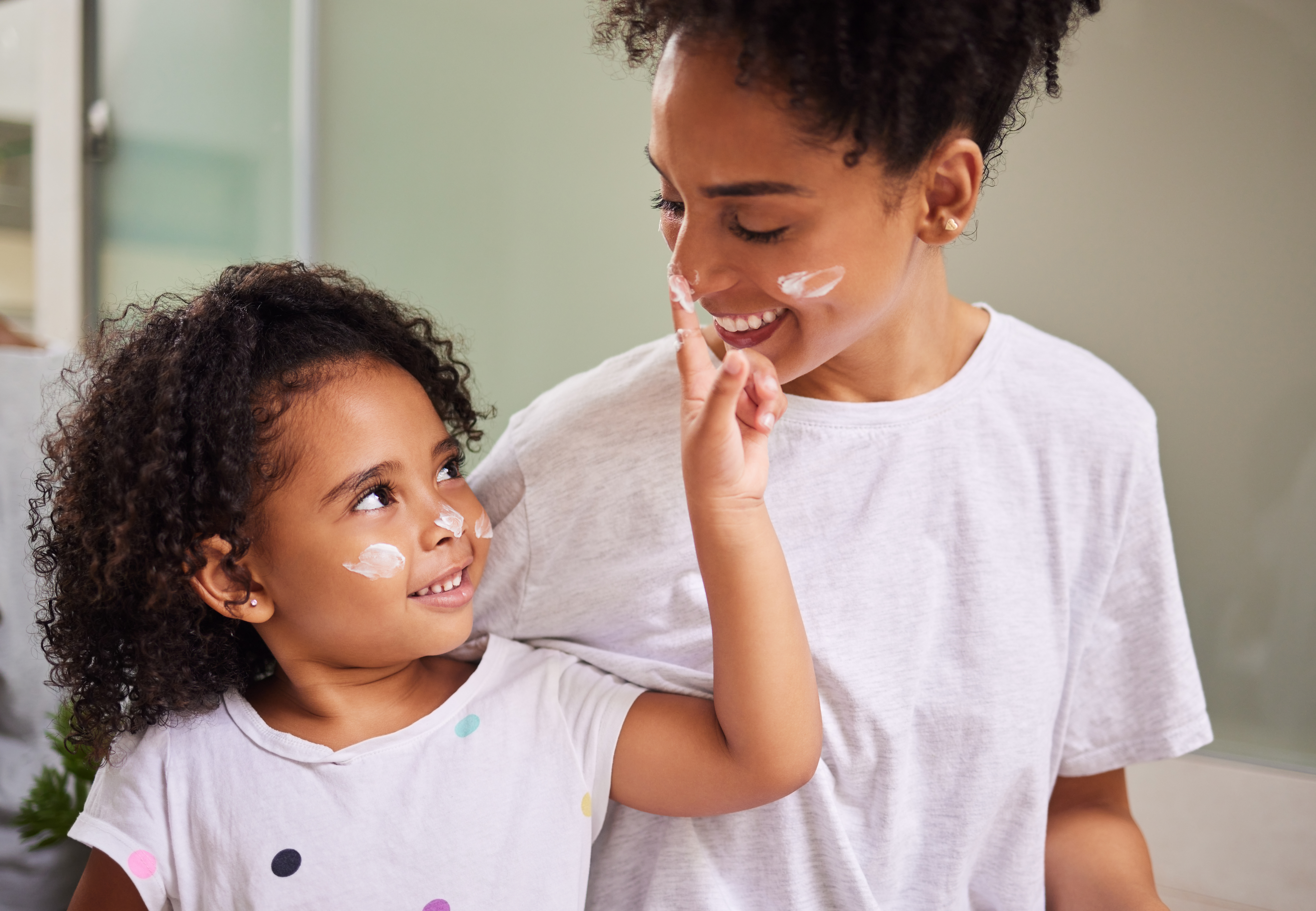 Happy family, mother and girl with cream skincare on face in the morning to bond in Mexico home. Smiling latino mom with young and cheerful child putting moisturiser on parent in house bathroom.