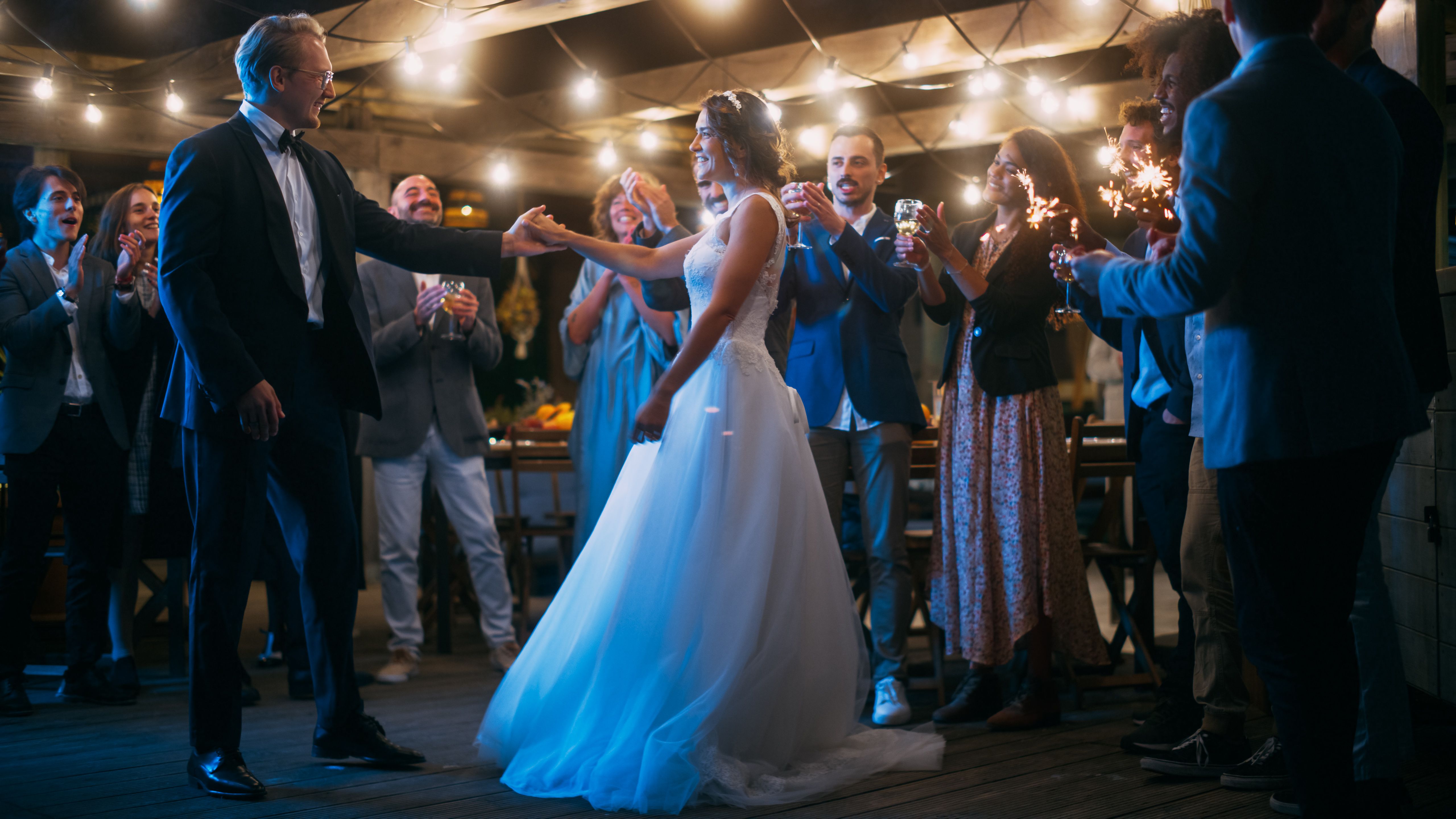Beautiful Bride in White Dress and Groom in Stylish Black Suit Celebrate Wedding at an Evening Reception Party. Newlyweds Dancing at a Venue with Best Multiethnic Diverse Friends.