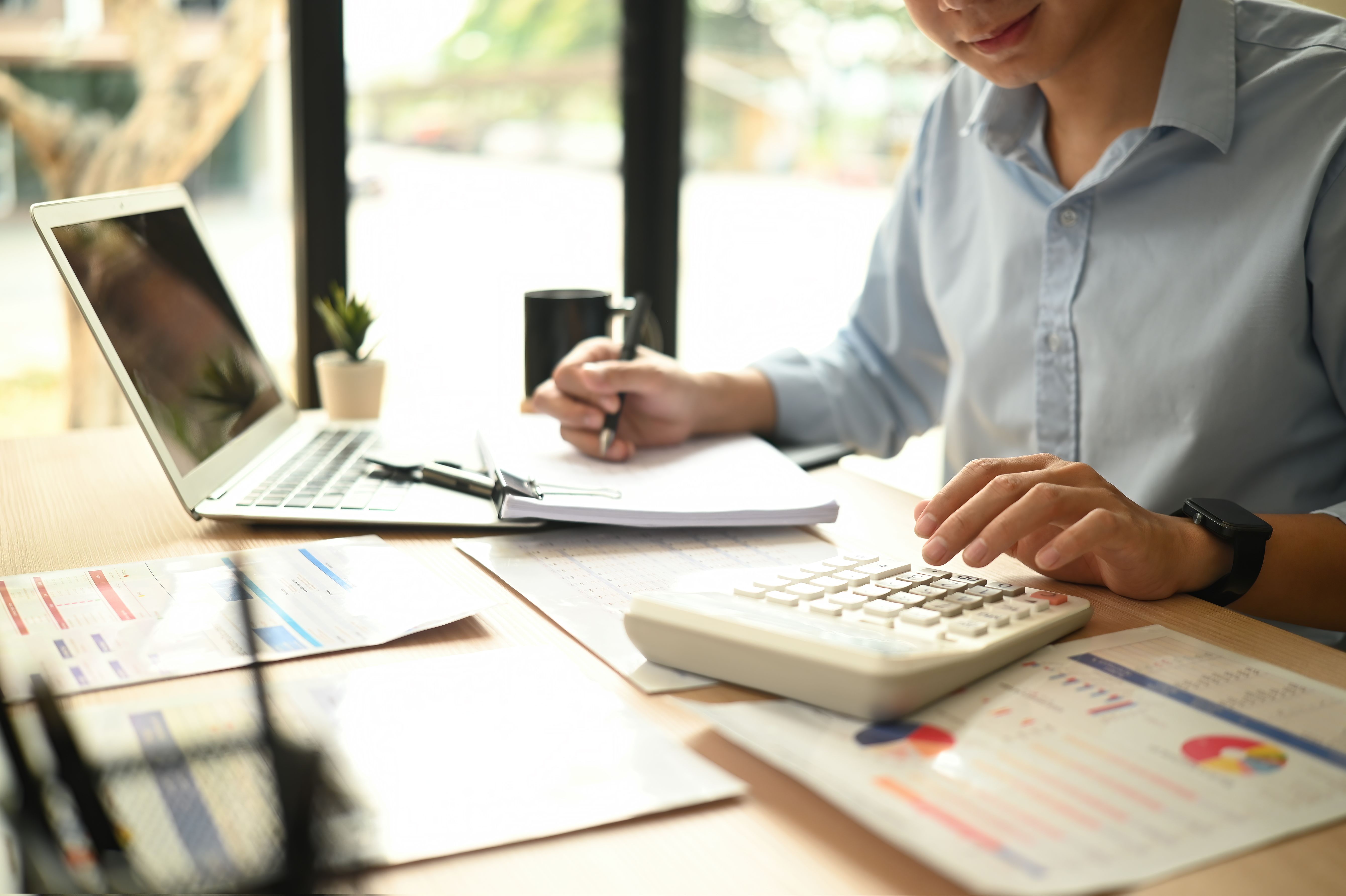 Cropped image of a Business analyst man using a calculator to evaluate financial charts and documents on a desk with a laptop and pen in hand